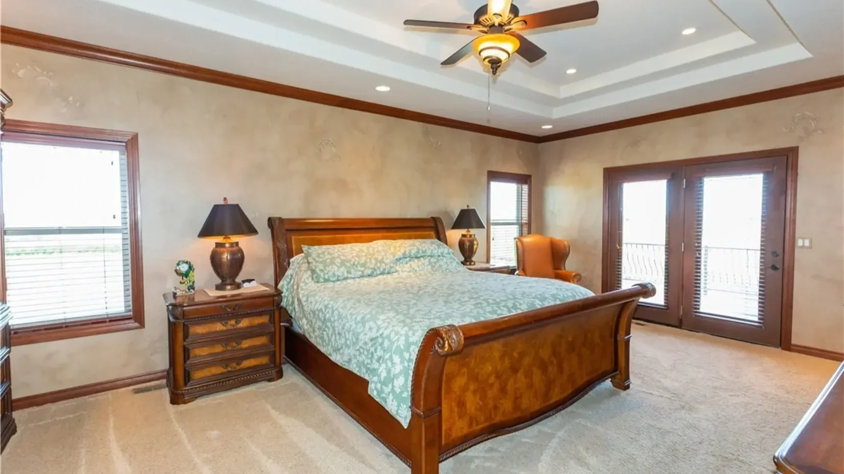Bedroom with wooden furniture, a bed with light patterned bedding, two bedside tables with lamps, an armchair, and large windows with blinds and a door to a balcony.