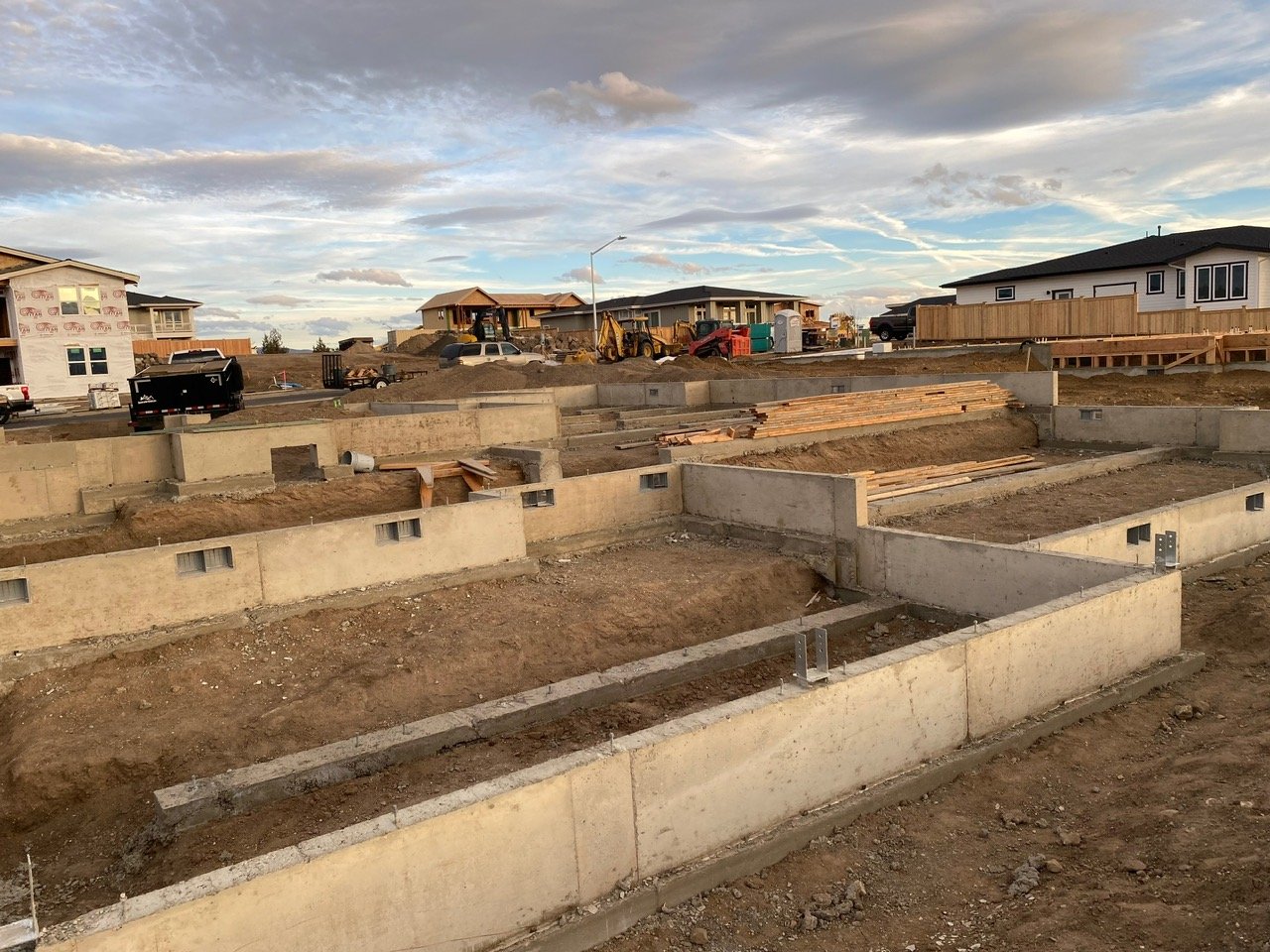 Construction site with building foundations and framing materials, in a residential area with houses, equipment, and a cloudy sky.