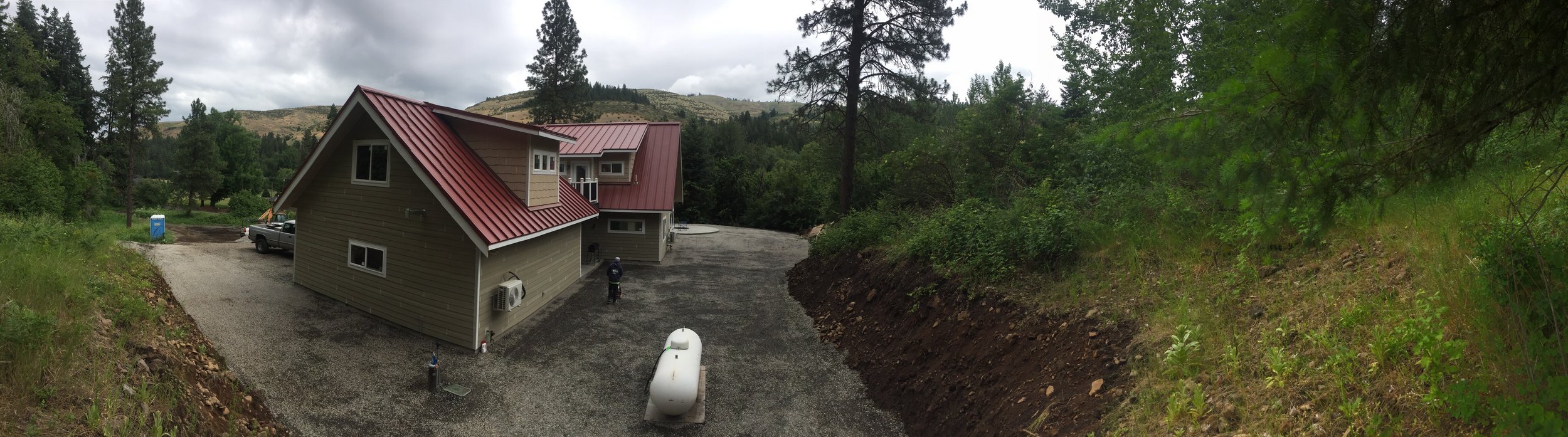 Newly constructed house with a red metal roof, located in a wooded area with trees and mountains in the background. There are vehicles, a portable toilet, and construction equipment visible near the house.