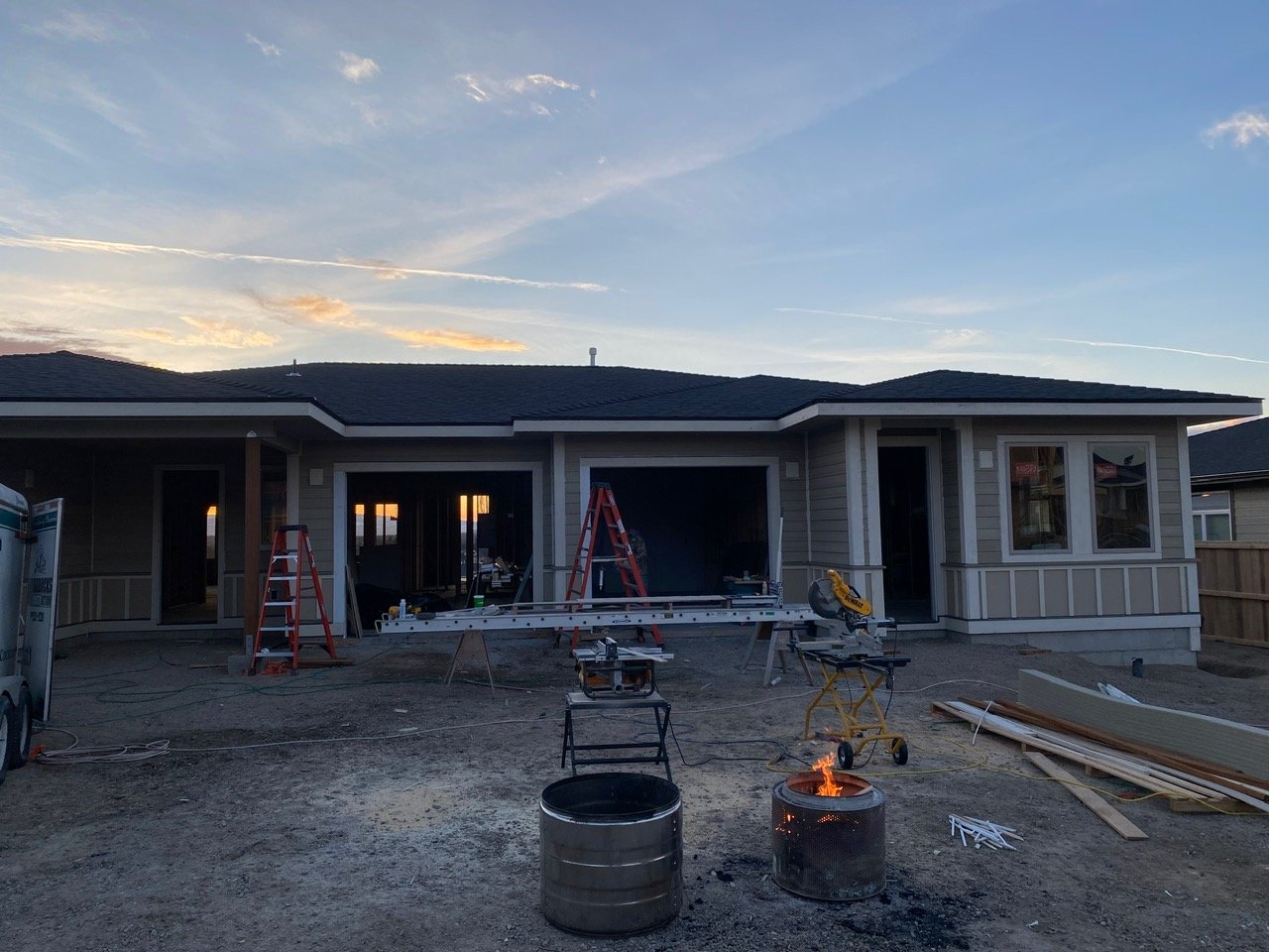 Under construction house with ladders, tools, and a small fire pit outside at sunset.