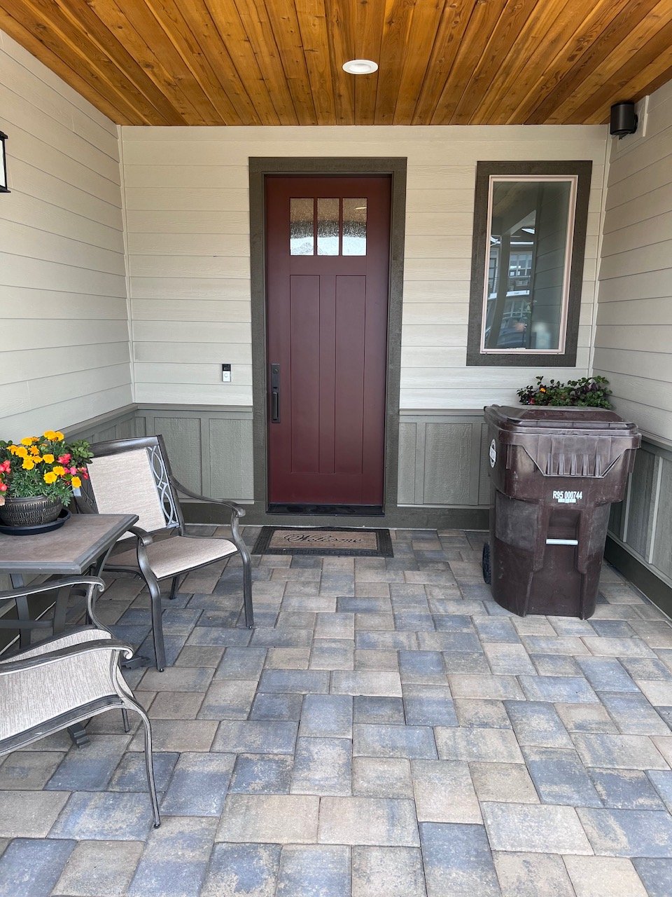 Covered porch area with a maroon front door, a window to the right, a trash bin, a small table with flowers, and two chairs with cushions, under a wooden ceiling.