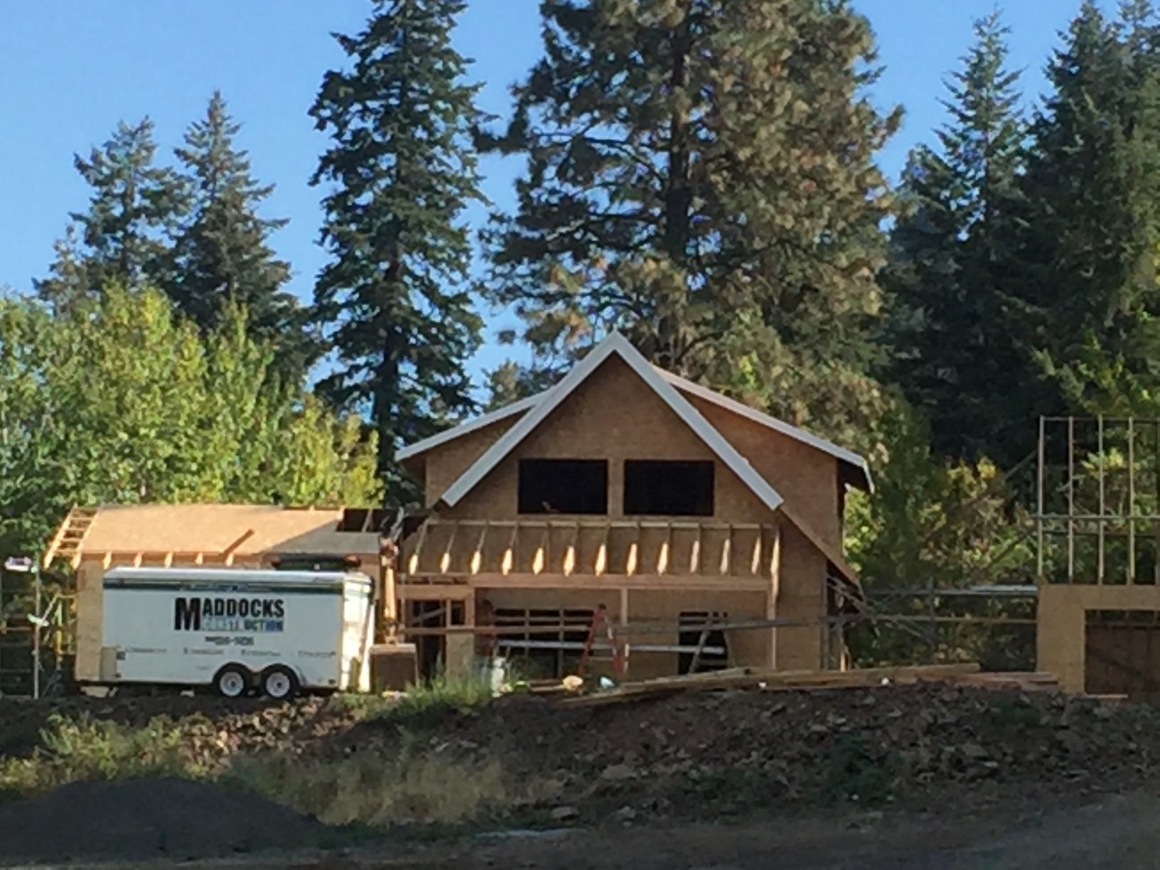 A house under construction with a wooden frame structure, surrounded by trees, a construction trailer labeled 'MADDOCKS ROOFING,' and construction materials.
