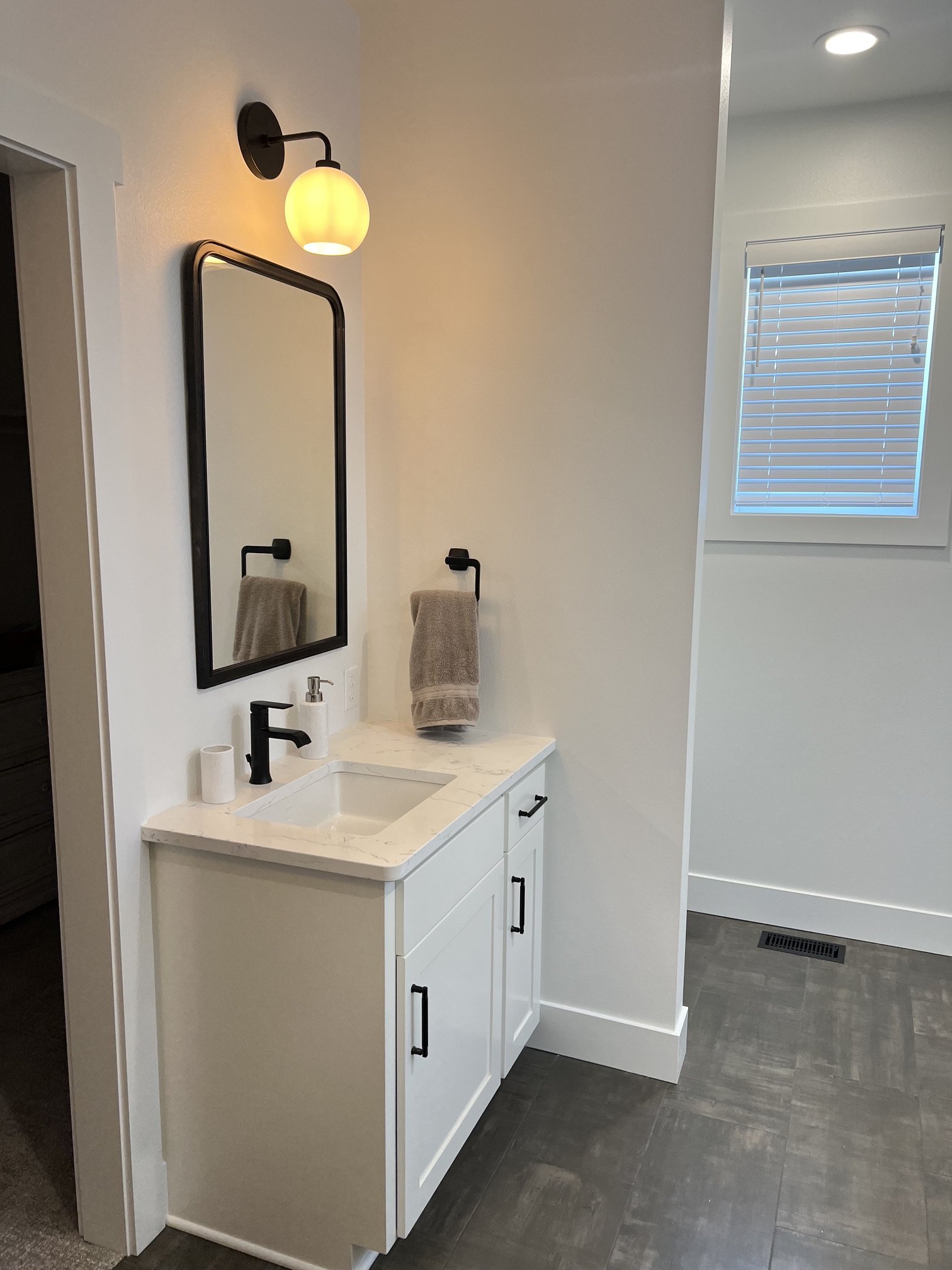 A small bathroom vanity with a black-framed mirror, a modern black faucet, a soap dispenser, and a towel on a towel ring, illuminated by a wall-mounted light fixture, with a window with blinds and gray tiled flooring.