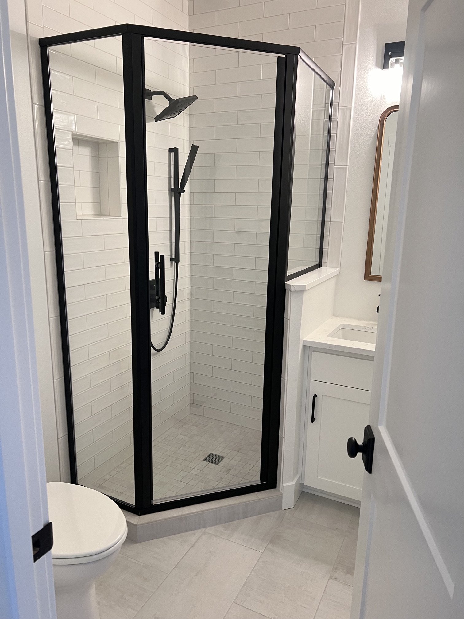A modern bathroom with a corner glass shower enclosure featuring white subway tile walls and a black metal frame, a toilet, and a white vanity with black hardware.