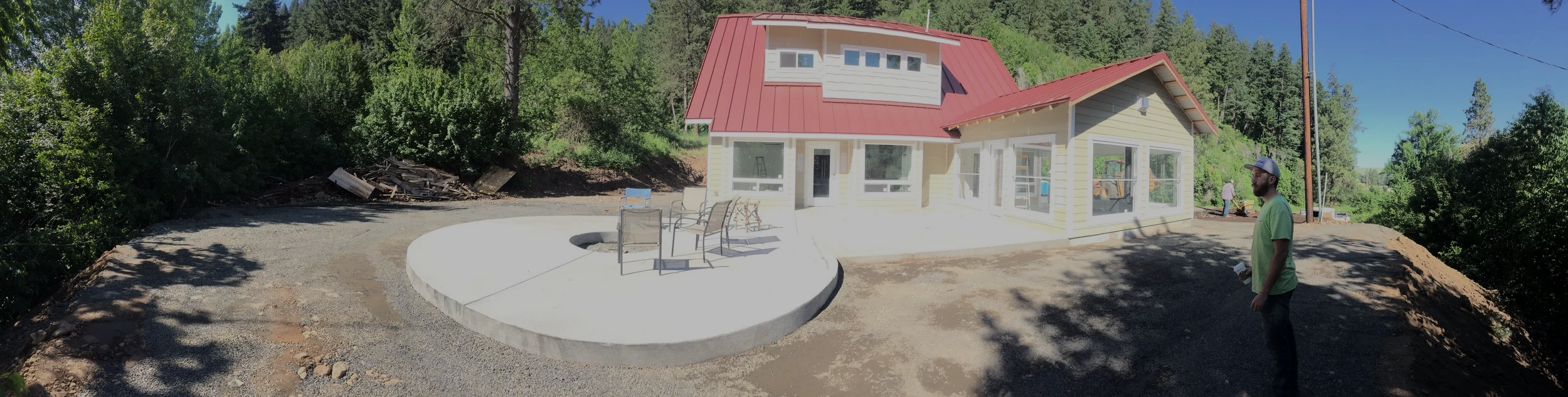 New yellow house with a red metal roof, large windows, and a concrete patio with outdoor furniture, surrounded by trees, with a man in a green shirt and glasses standing nearby.