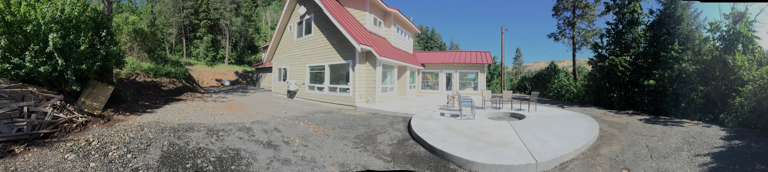 A newly constructed house with a red metal roof, beige siding, and white trim, situated on a cleared lot with gravel and dirt, surrounded by trees and blue sky.