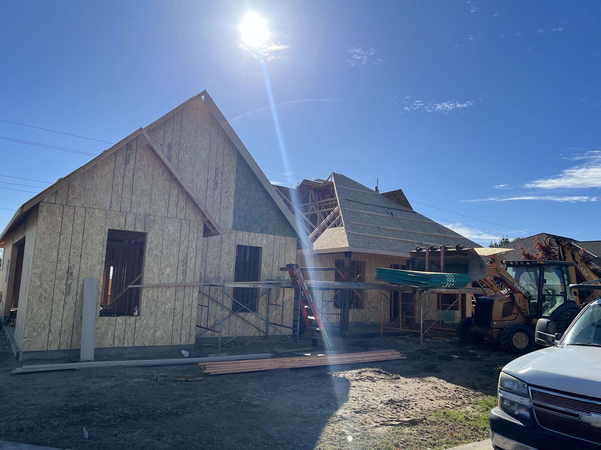 A house under construction with wooden framing and sheathing, a construction crane, and construction vehicles. The sky is clear with sunlight shining.