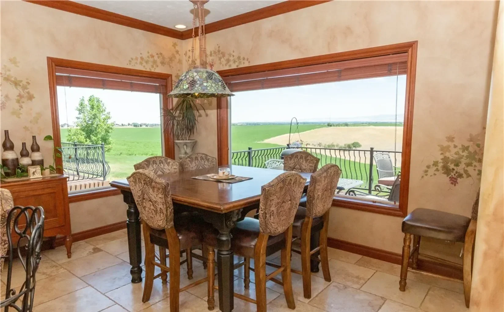 Dining room with a large wooden table surrounded by six ornate upholstered chairs, large windows with views of green fields and a balcony, and a decorative chandelier overhead.