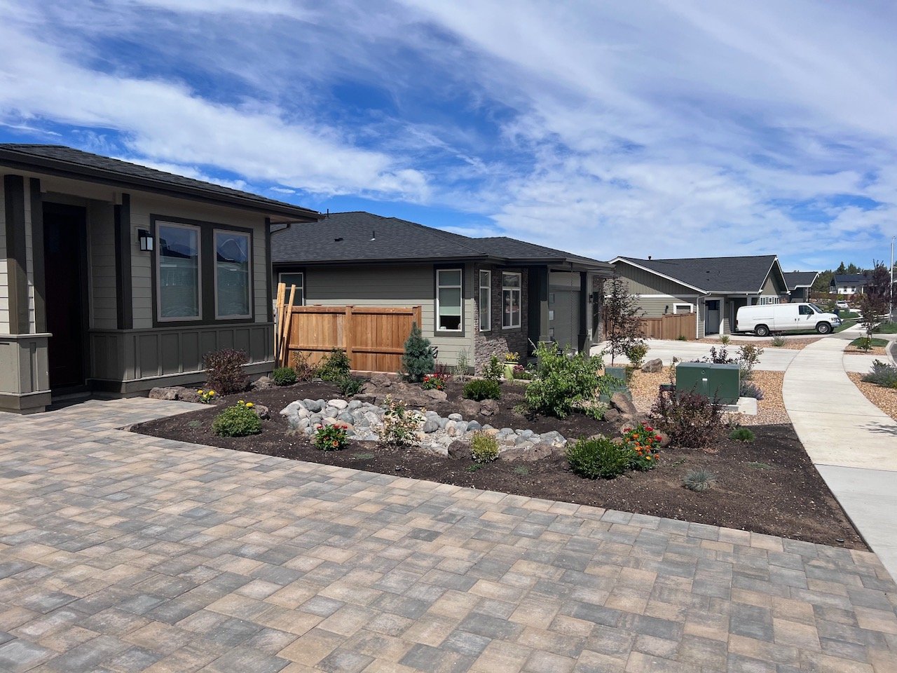 Front yard of a suburban house with a brick driveway, landscaped garden beds, and a curved sidewalk under a partly cloudy sky.