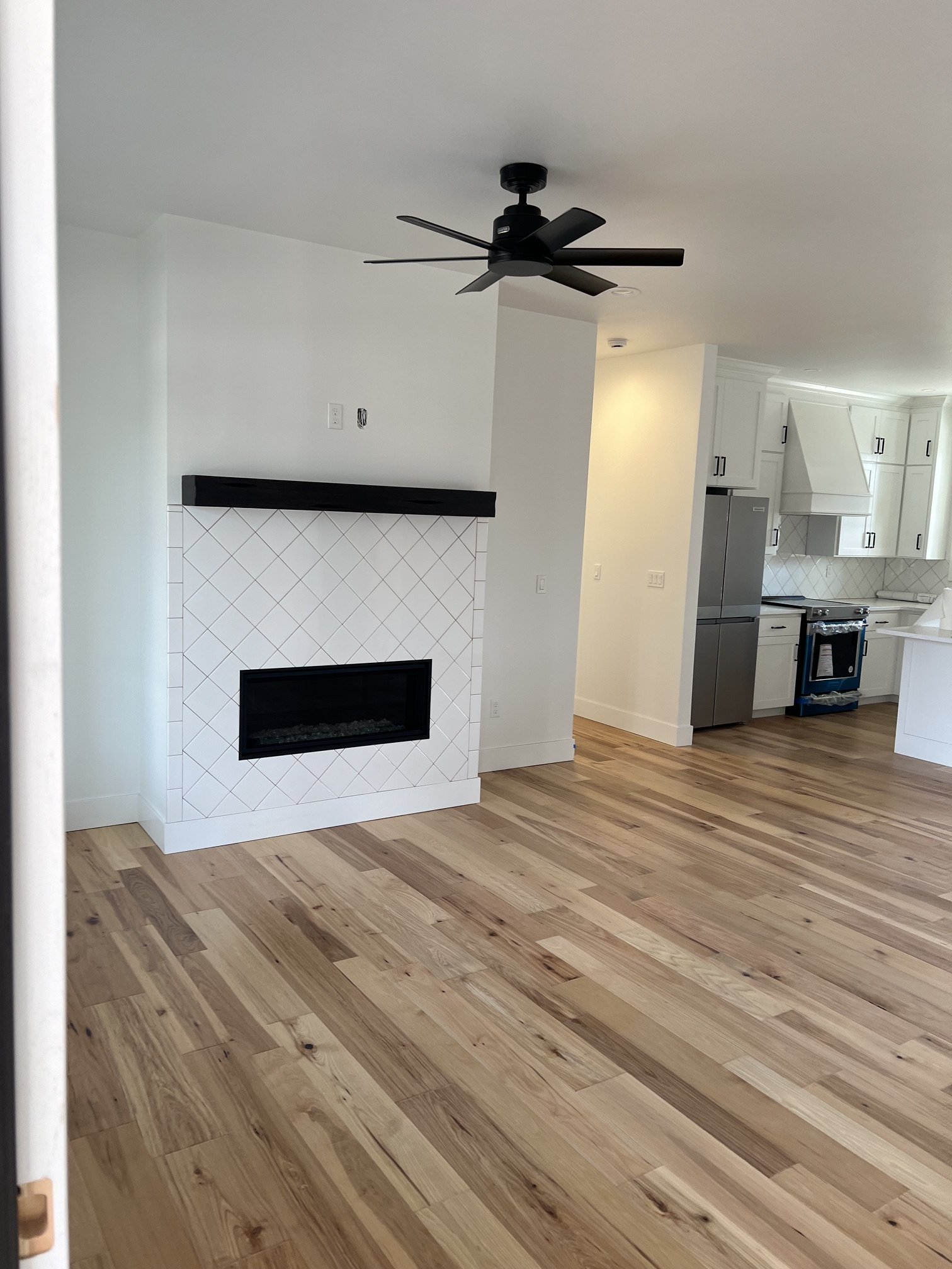 Living room with hardwood flooring, white walls, a black ceiling fan, and a modern white fireplace with a black trim, adjacent to a kitchen area with white cabinets and stainless steel appliances.