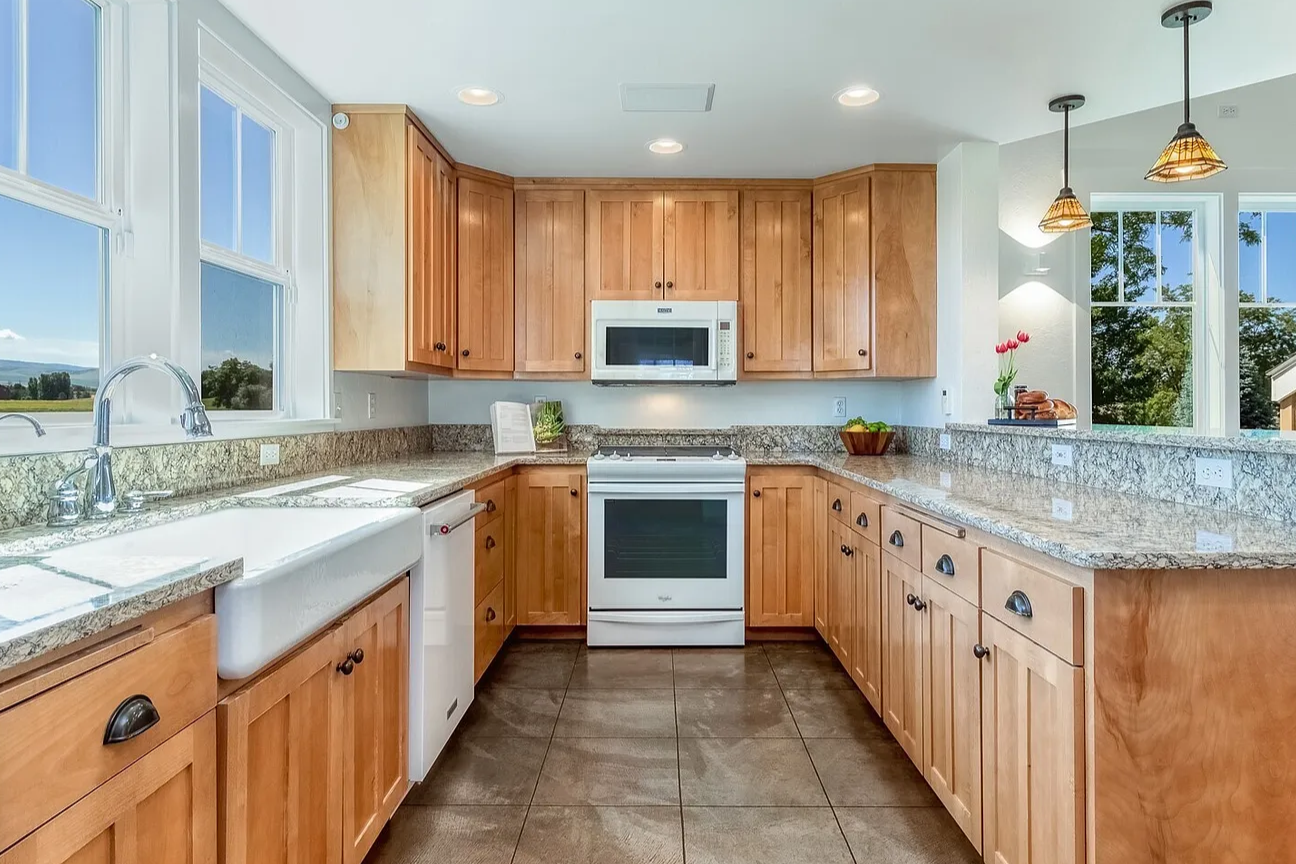 A kitchen with wooden cabinets, granite countertops, a white sink, and a white stove with a microwave above. There are large windows with a view of trees and a blue sky.