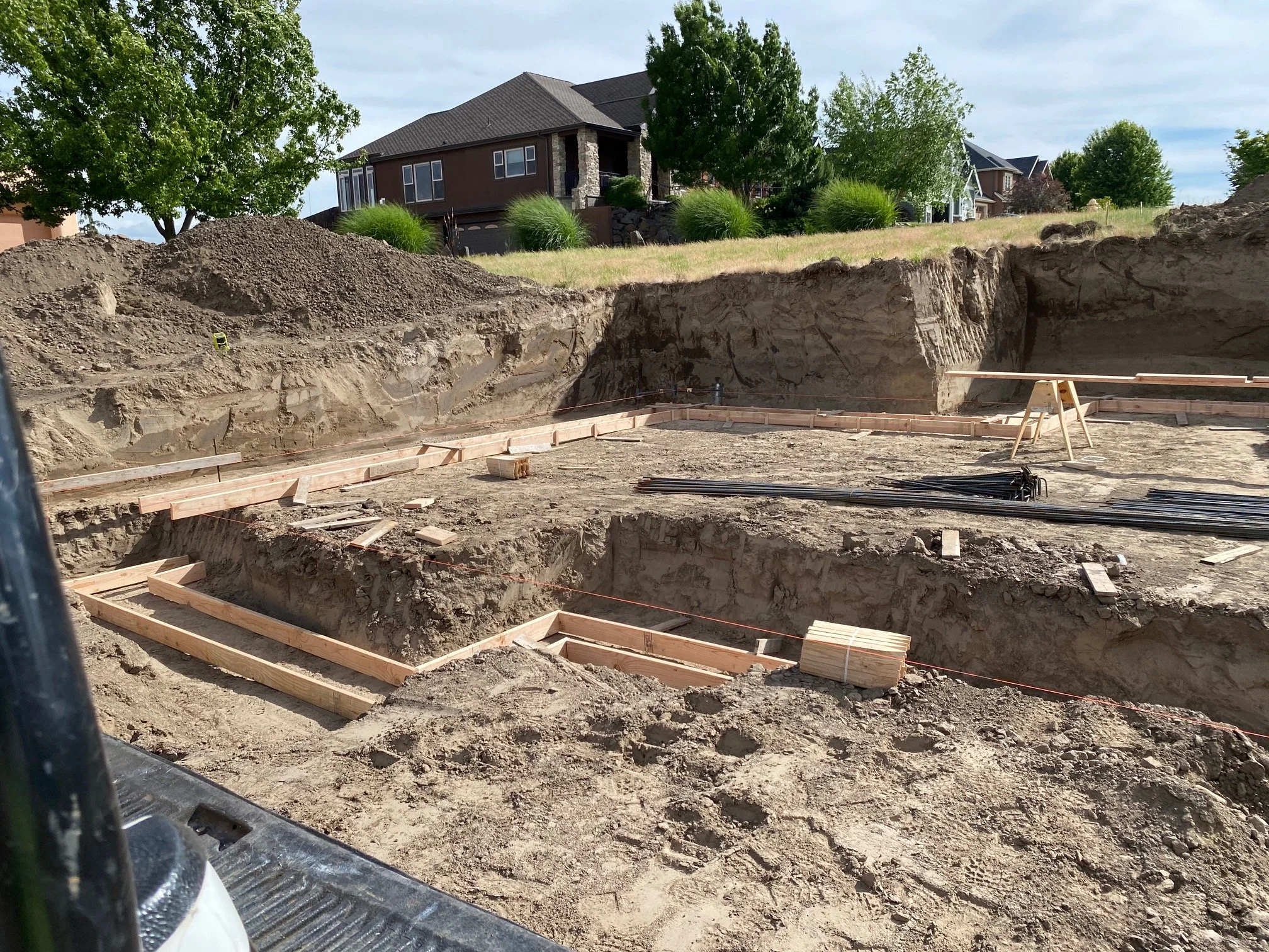 Construction site with an excavated trench in the foreground, wooden forms for concrete foundation, and residential houses with trees in the background.