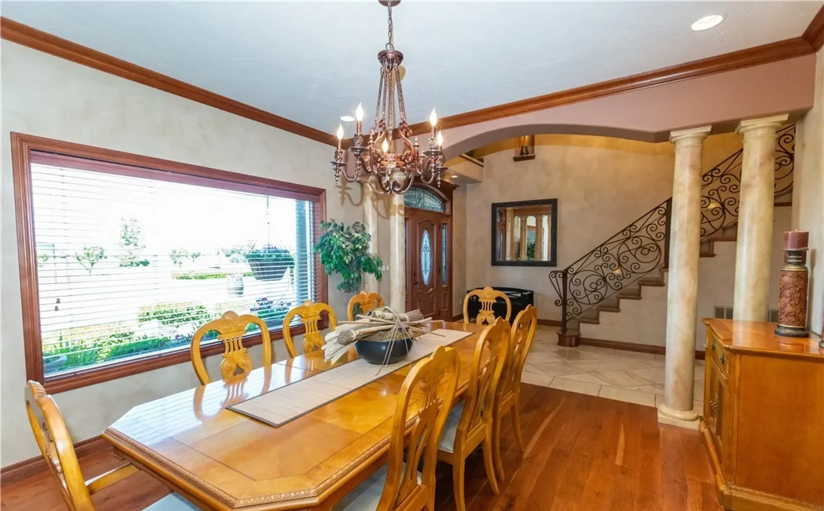 Dining room with large wooden table, eight chairs, chandelier, window with blinds, and decorative columns near staircase.