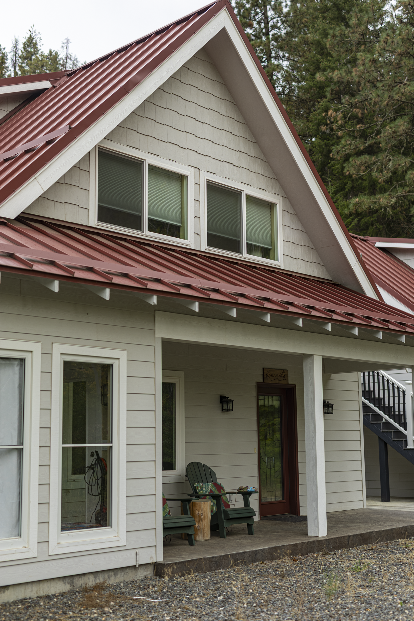 View of a modern house with beige siding, a red metal roof, and a porch area with outdoor chairs. The house has large windows and is surrounded by trees.