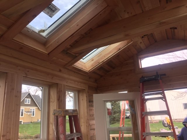 Interior of a room under construction with wooden walls, two skylights, and three windows; ladders inside.