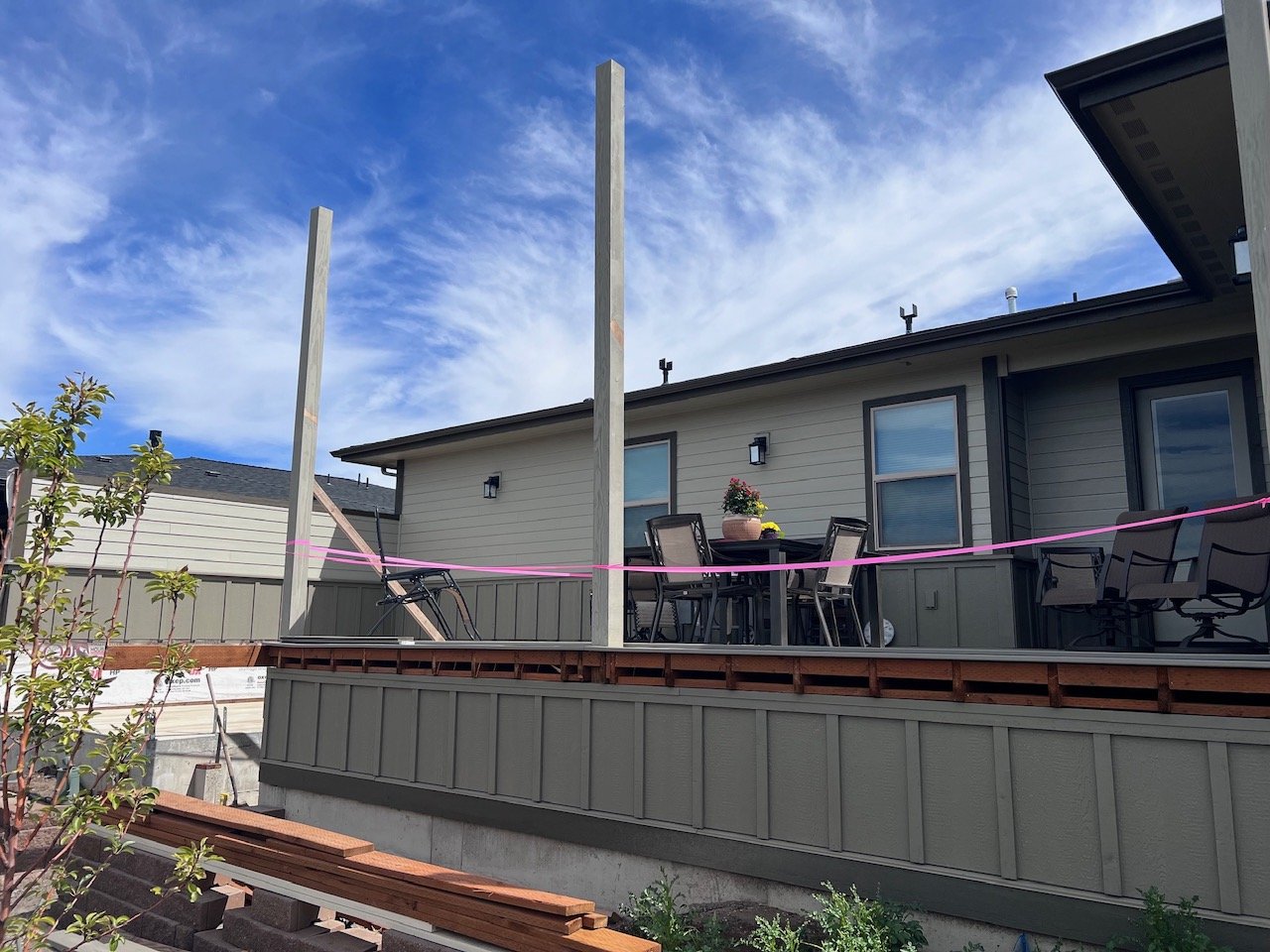 Backyard deck with chairs and table under construction, with wooden posts and a pink construction ribbon, against a bright blue sky with clouds.