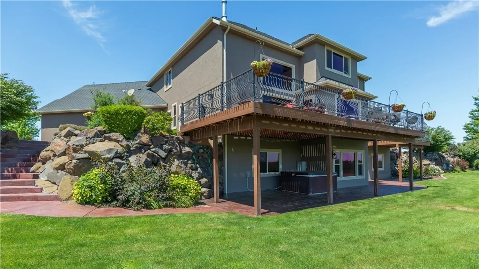 A two-story house with a large wooden deck on the upper level, decorated with hanging flower baskets. There are stairs on the left side leading up to the deck, and the house is surrounded by a well-maintained lawn and landscaped rocks with bushes. Th