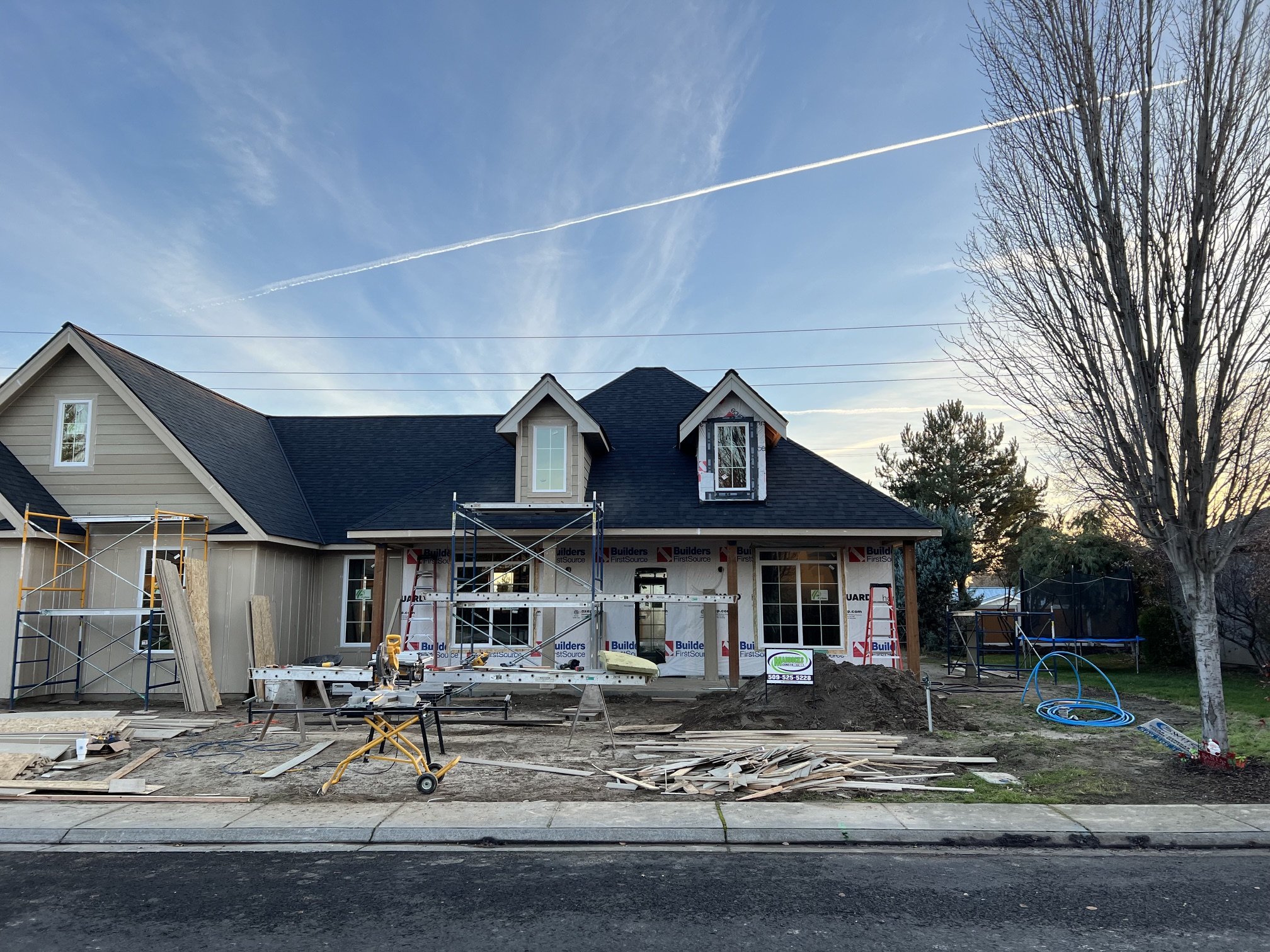 A house under construction with scaffolding and construction materials in the front yard, clear blue sky, and trees.