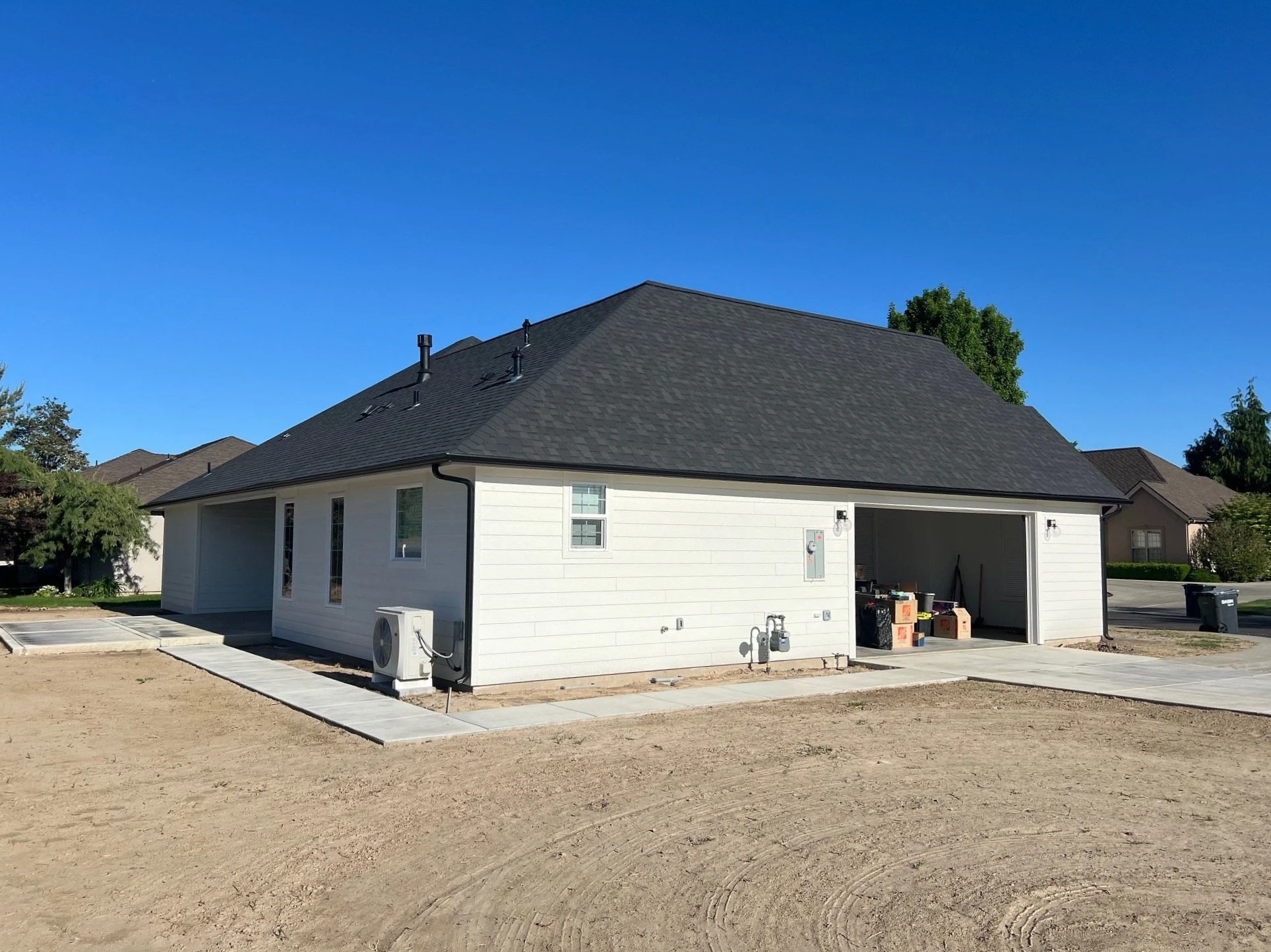 A newly built white house with a dark gray roof and an open garage door. Construction materials and boxes are visible inside the garage. There are several vents on the roof, and an air conditioning unit is installed outside the house. The area around the house is under development, with bare dirt and a concrete sidewalk in front.