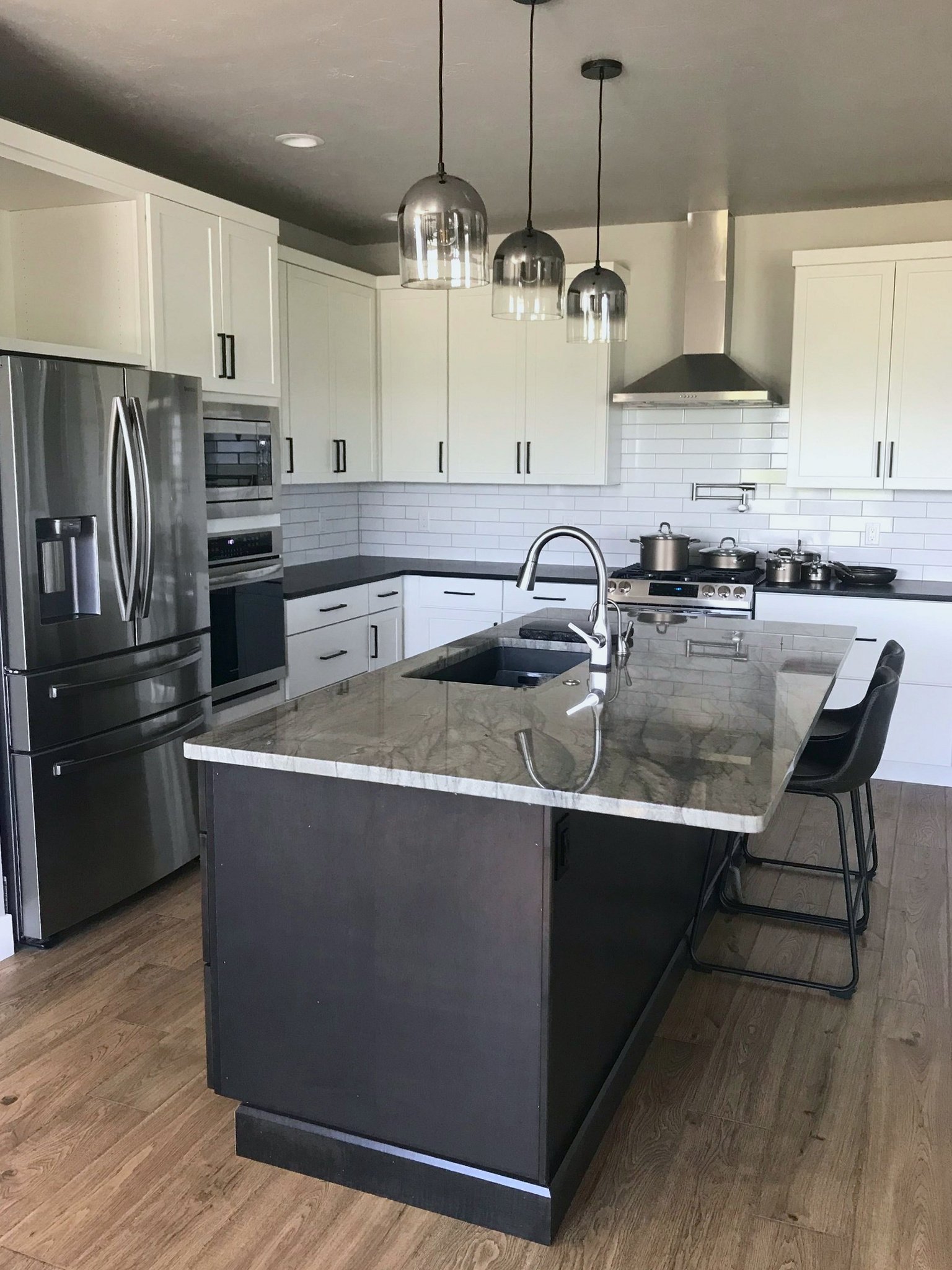 Modern kitchen with white cabinets, a black island with a granite countertop, stainless steel appliances including a refrigerator and an oven, pendant lights hanging over the island, a stainless steel range hood, and a hardwood floor.