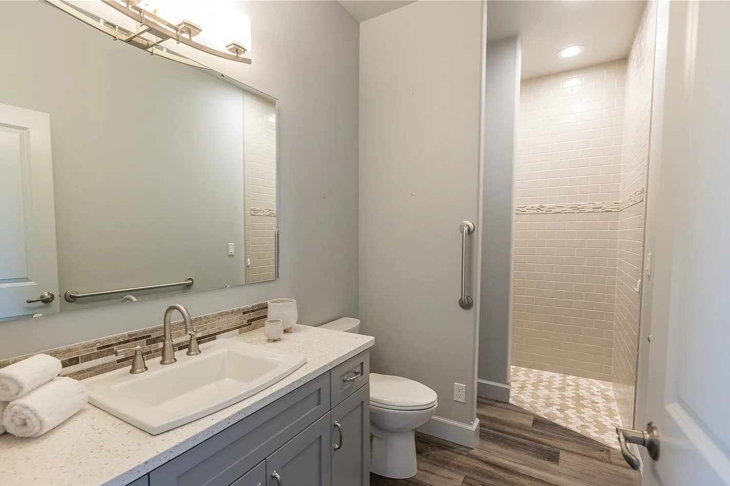 Modern bathroom with a large mirror, white sink, gray cabinetry, and a walk-in shower with white brick tiles and a decorative band, wood-look flooring.