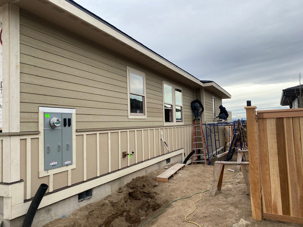 Construction workers installing siding on a beige house exterior with a fenced yard, windows, and building tools visible.