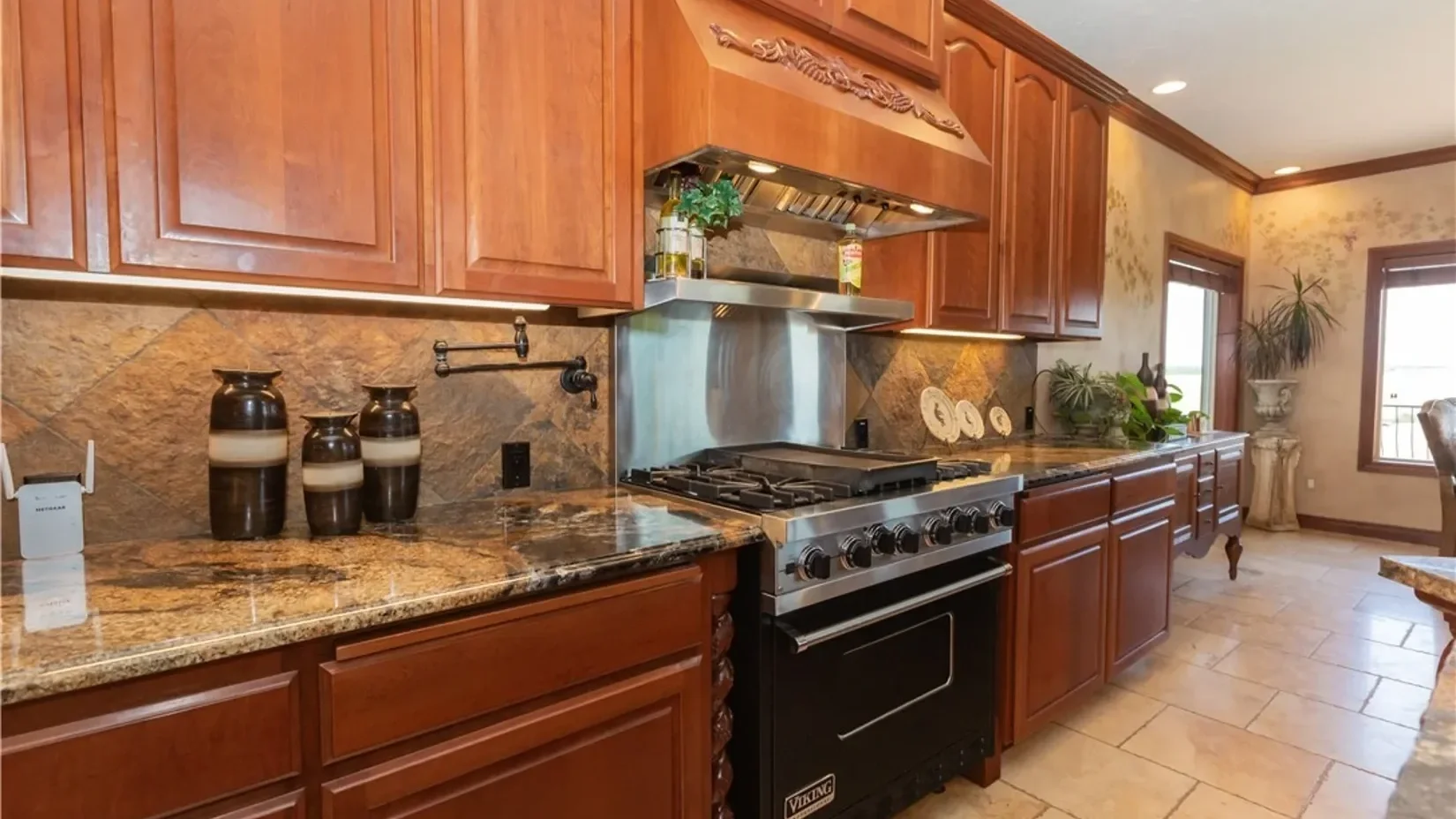 A modern kitchen with wooden cabinets, granite countertops, a stainless steel oven and range, decorative vases, plants, window blinds, and a tiled floor.