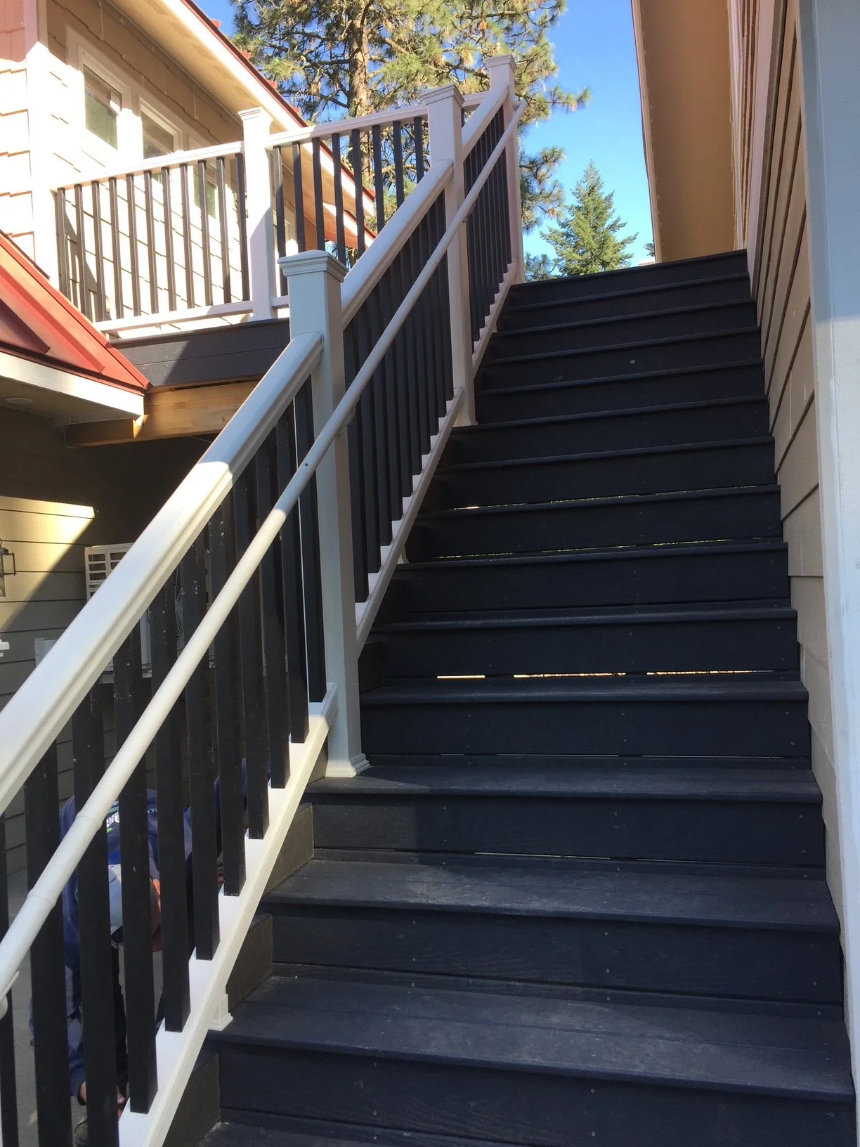 Exterior wooden staircase with black steps, white railings, and vertical black balusters, leading to a second-story balcony of a house. The house has beige siding, and there are tall trees and a blue sky in the background.
