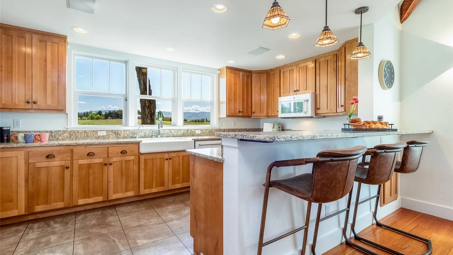 A bright kitchen with wooden cabinets, granite countertops, a white farmhouse sink, and three brown leather bar stools at a breakfast bar. Large windows reveal a green field and trees outside, with a mountain landscape in the distance.