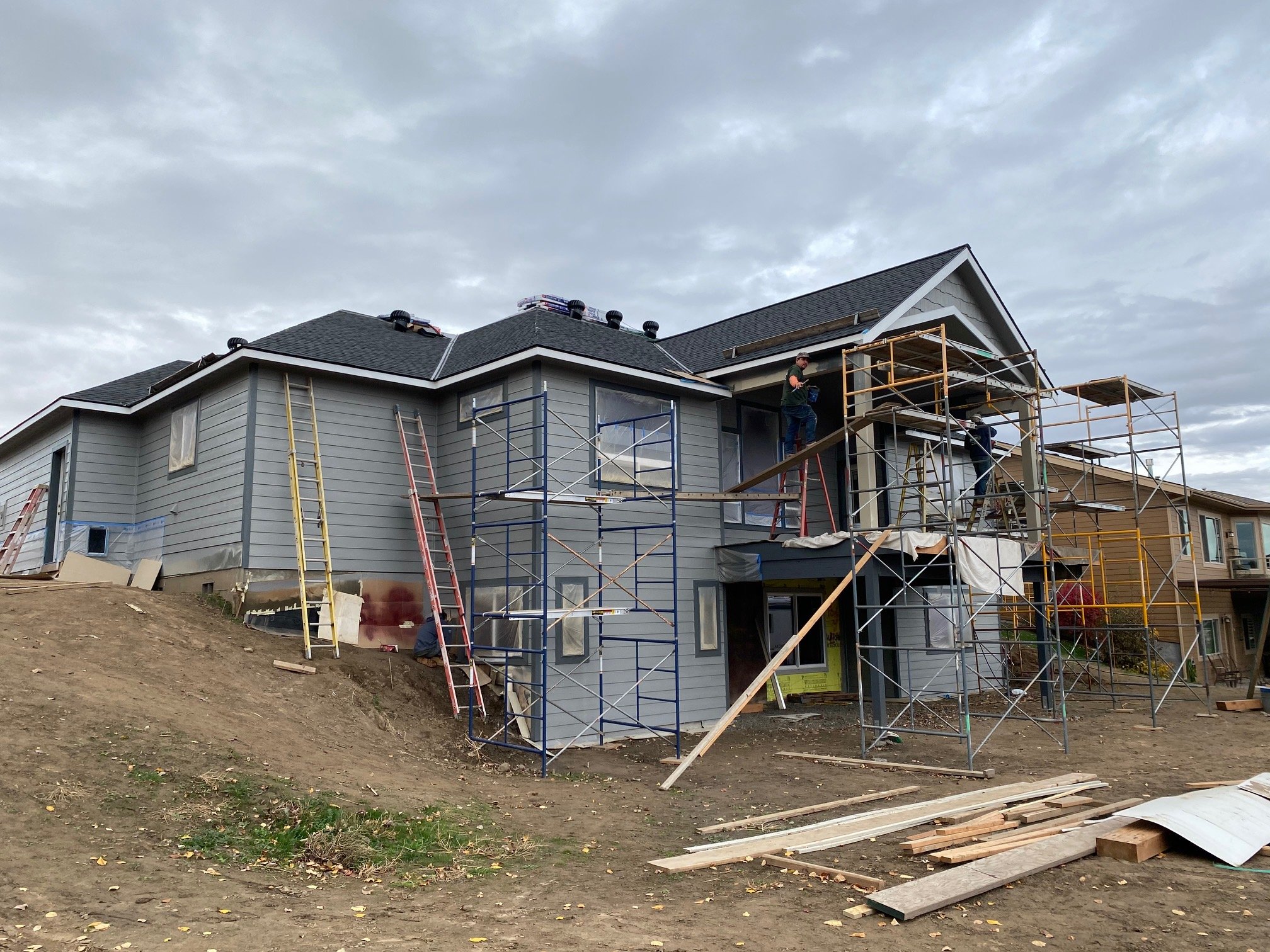 A house under construction with scaffolding around it, workers installing siding or roofing, and an overcast sky.
