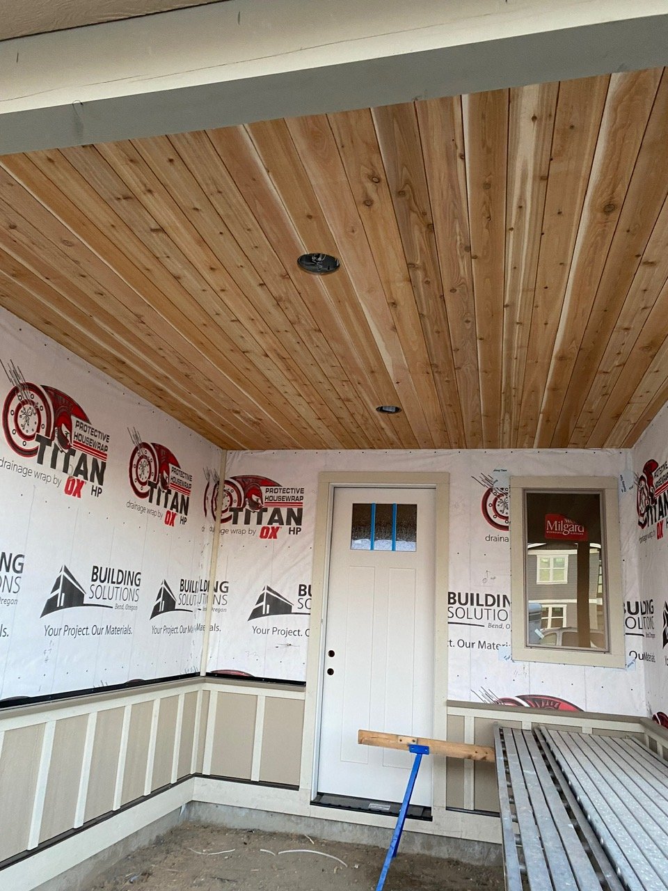 Interior under construction with a wooden plank ceiling, a white door with small window, window opening, and protective house wrap on the walls.