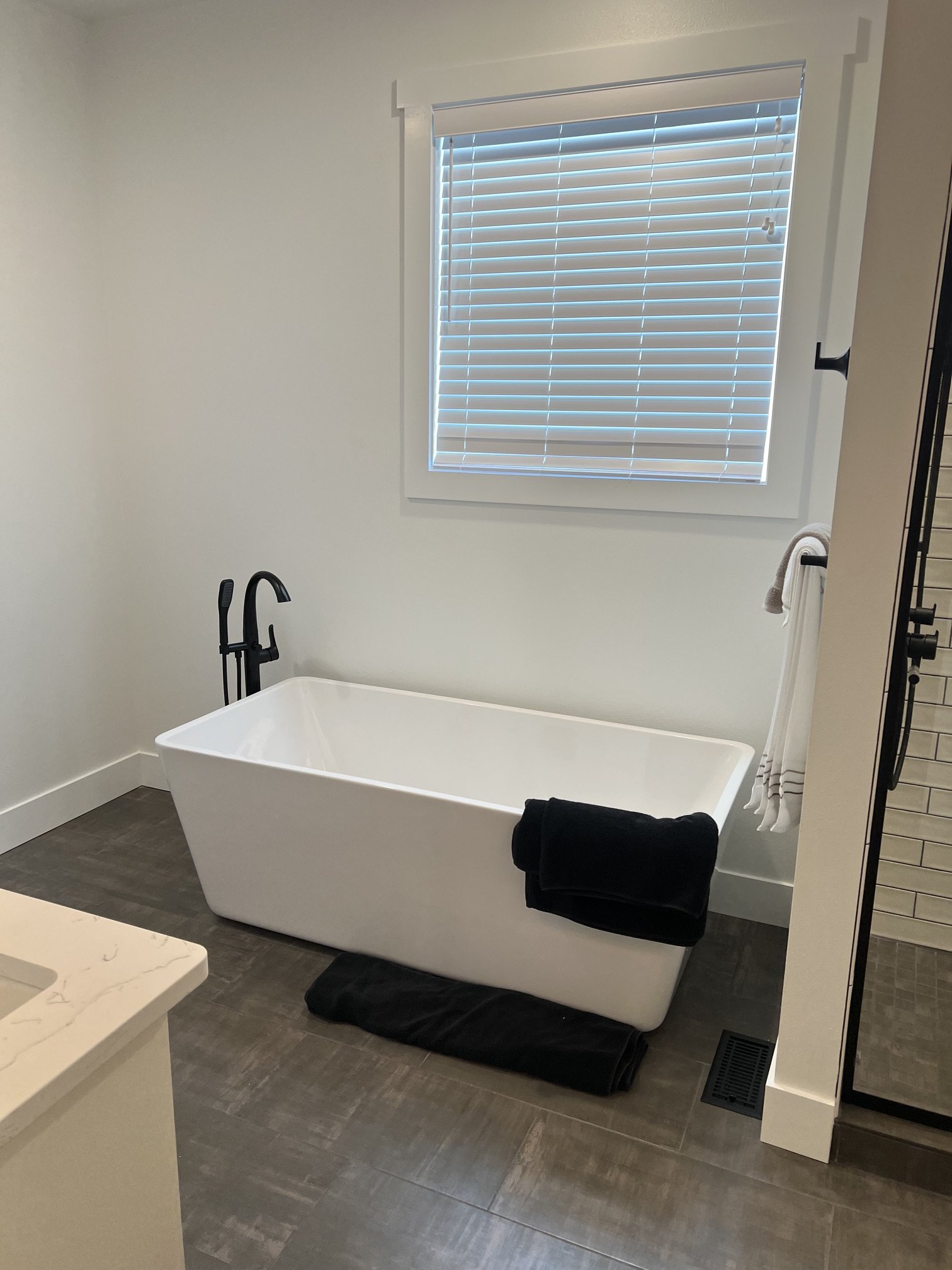 Bathroom with freestanding white bathtub below a window with closed white blinds, black towels on the edge of the bathtub, a black faucet, and a towel bar with a white towel inside a modern bathroom with gray tiled floor.