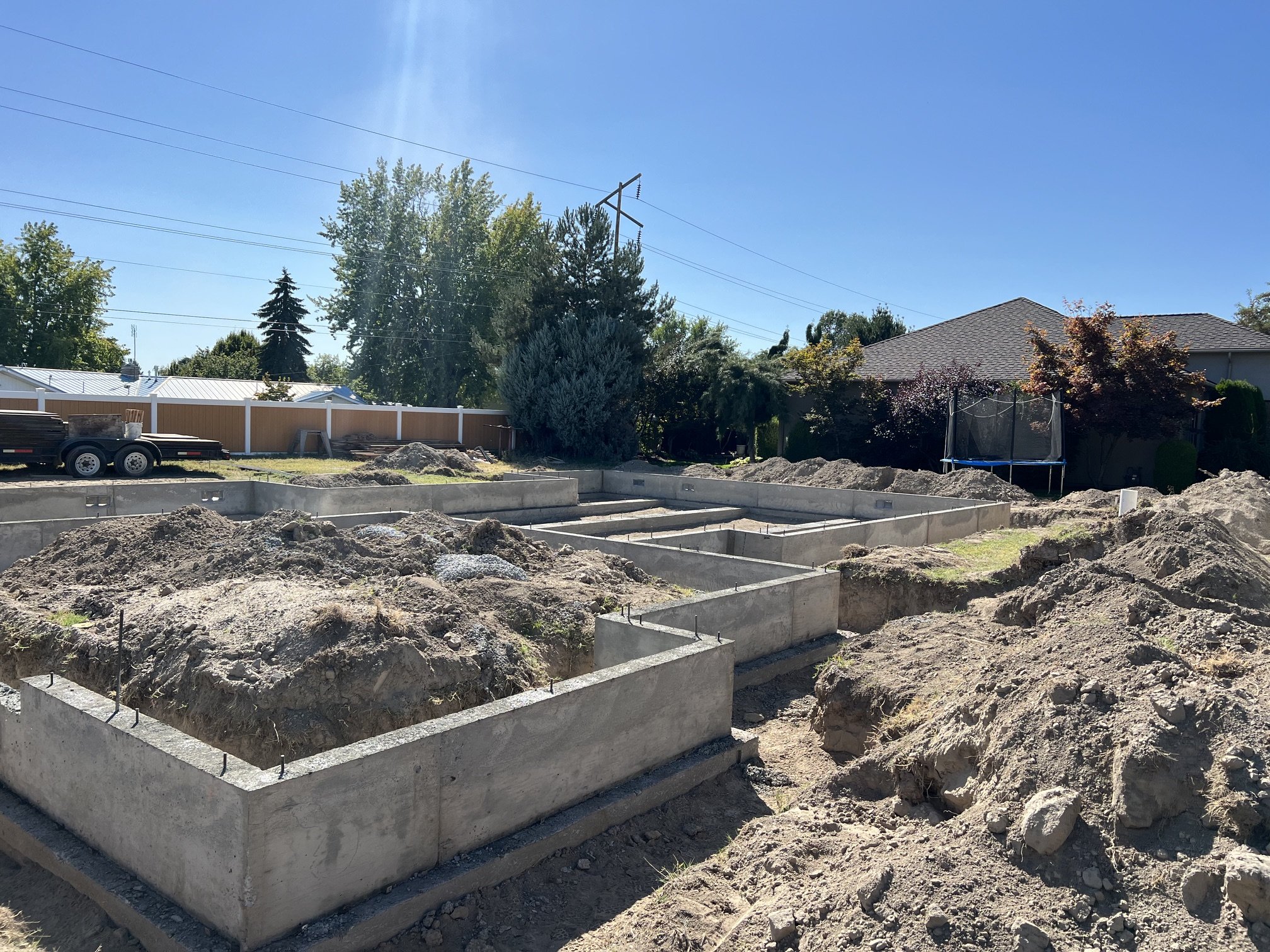 Construction site with concrete foundation outlines and piles of dirt, in a residential area with trees and houses in the background.