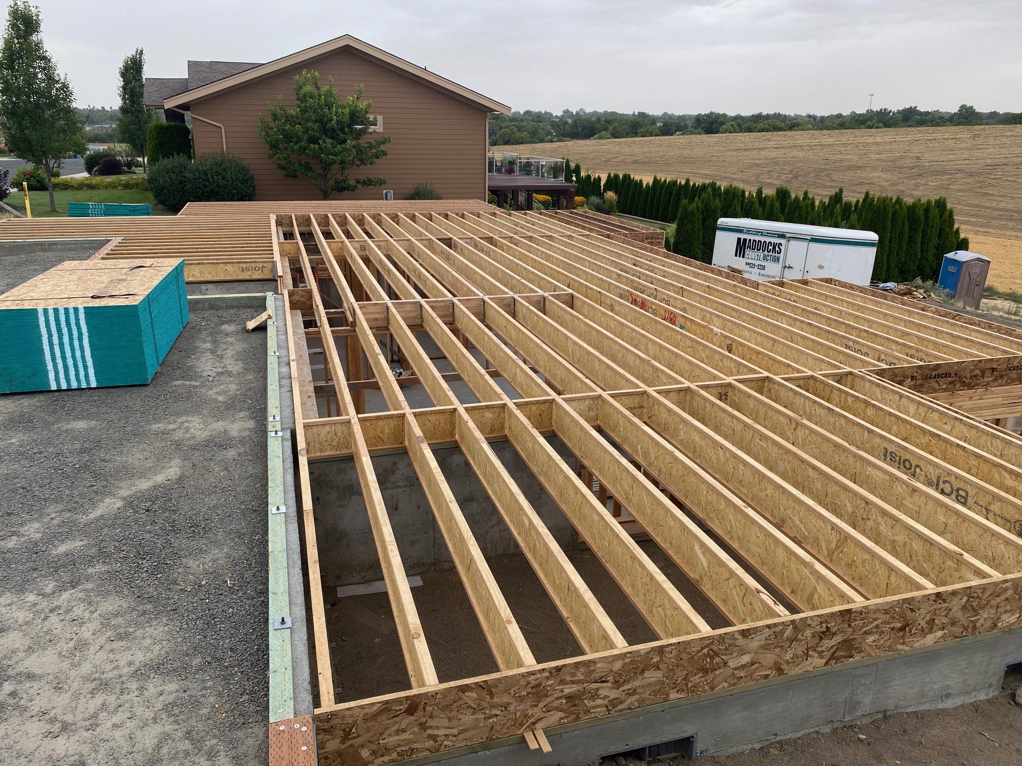 Construction site with wooden framing for a building, construction materials, and equipment in the background, including a Maddocks Construction trailer and portable toilet.