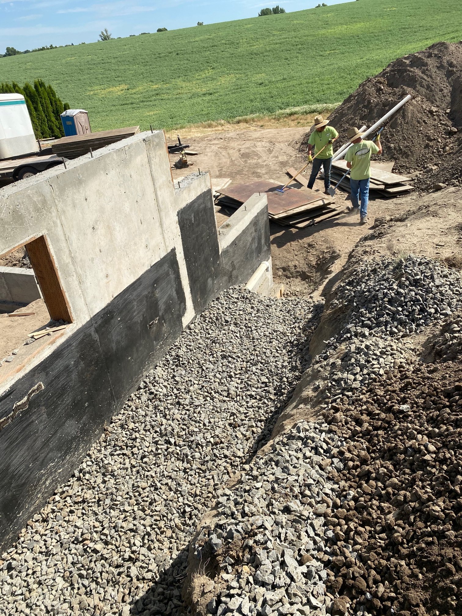 Two construction workers wearing hats and neon green shirts working on a construction site, spreading gravel on the ground, with construction materials and pipes nearby, and a green landscape and blue sky in the background.