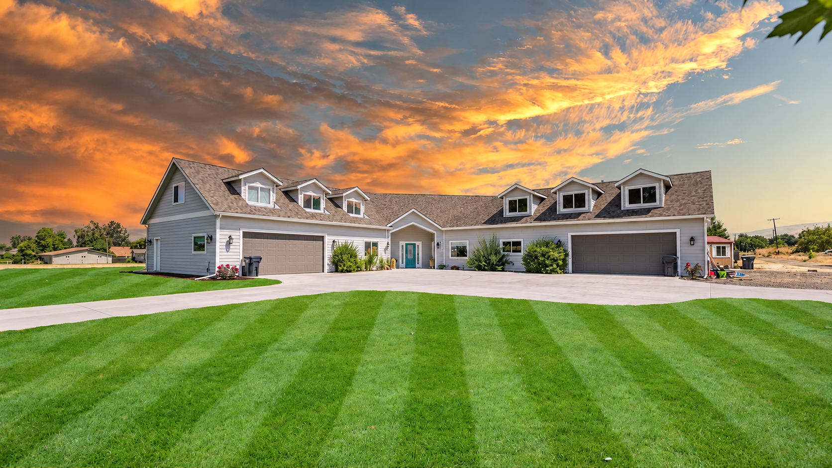 Front view of a large modern house with a spacious driveway, well-maintained green lawn, and trees, against a vibrant sunset sky with orange and yellow clouds.