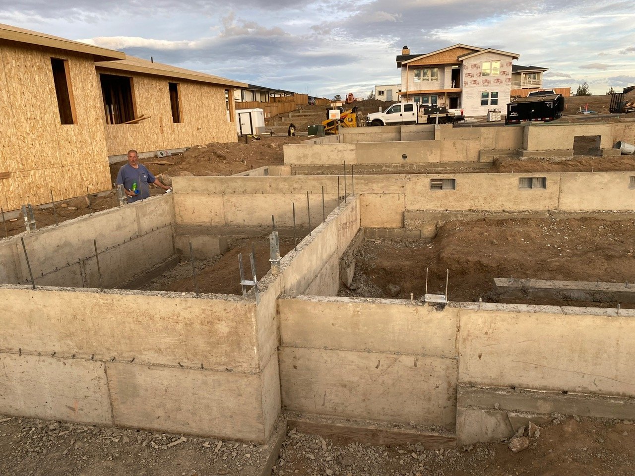 Construction site with concrete foundations and partially built houses, with a man holding a drink in the foreground and equipment and vehicles in the background.