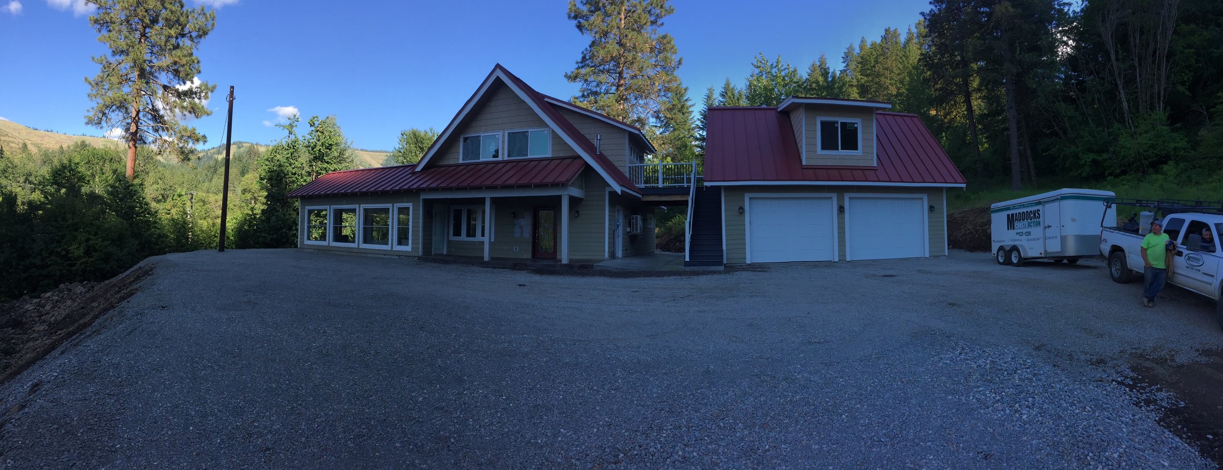 A newly constructed house with a red metal roof, multiple large windows, and a garage with two doors, situated in a green mountainous area with tall trees. Two men are standing near a white pickup truck and enclosed trailer on a gravel driveway.
