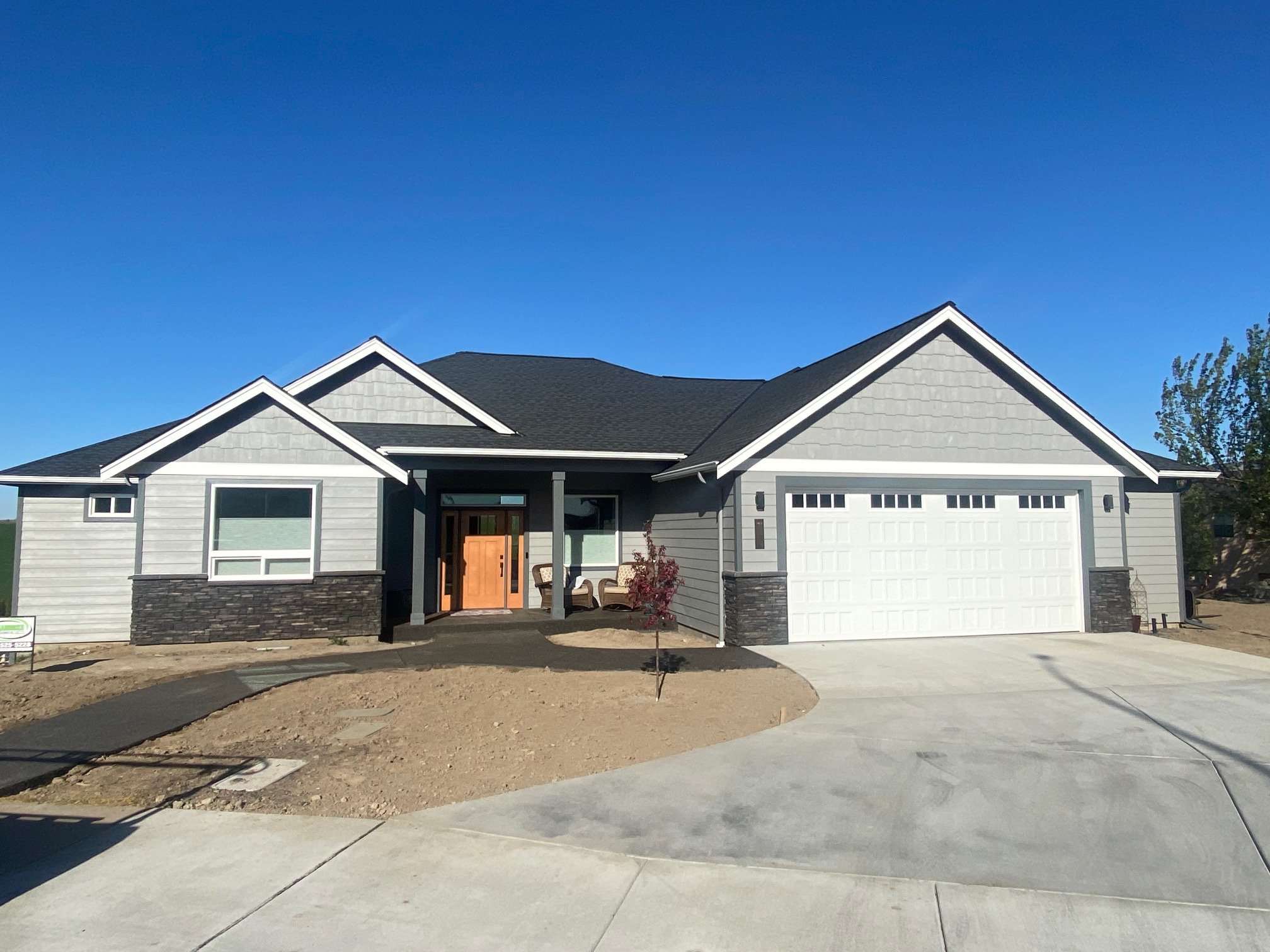 Newly constructed house with a grey exterior, white garage door, and a front porch with chairs, against a bright blue sky and minimal landscaping.