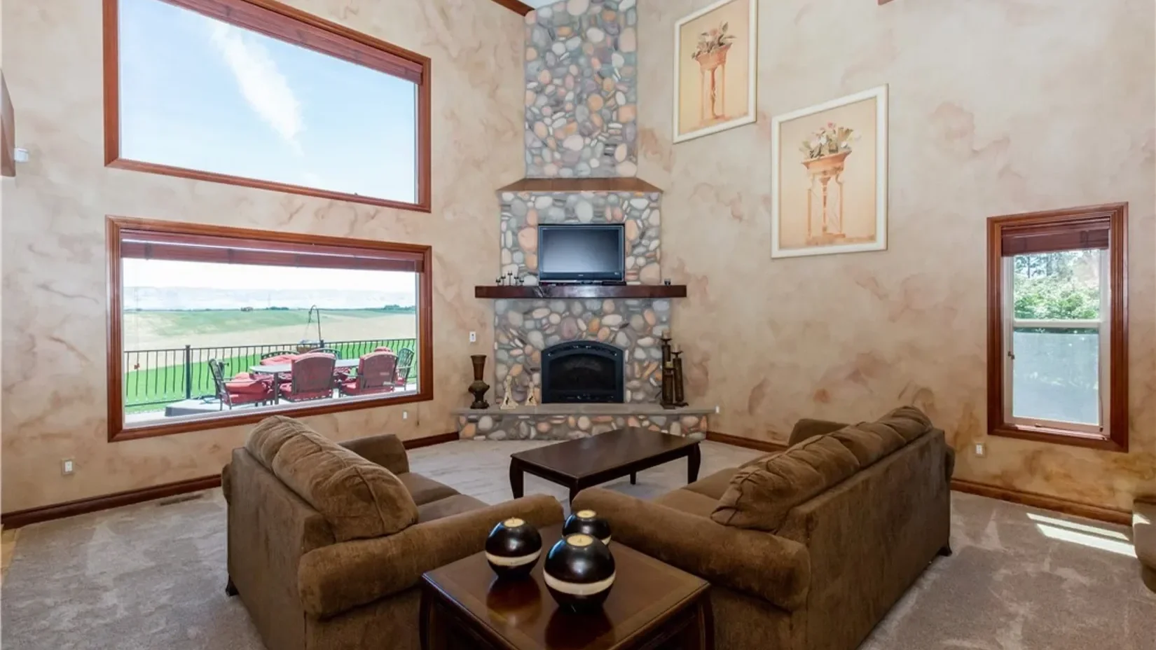 Living room with large windows showing a porch and green fields, a stone fireplace with a TV on mantel, two framed floral prints, two brown sofas around a coffee table, and a small table with three black and white vases.