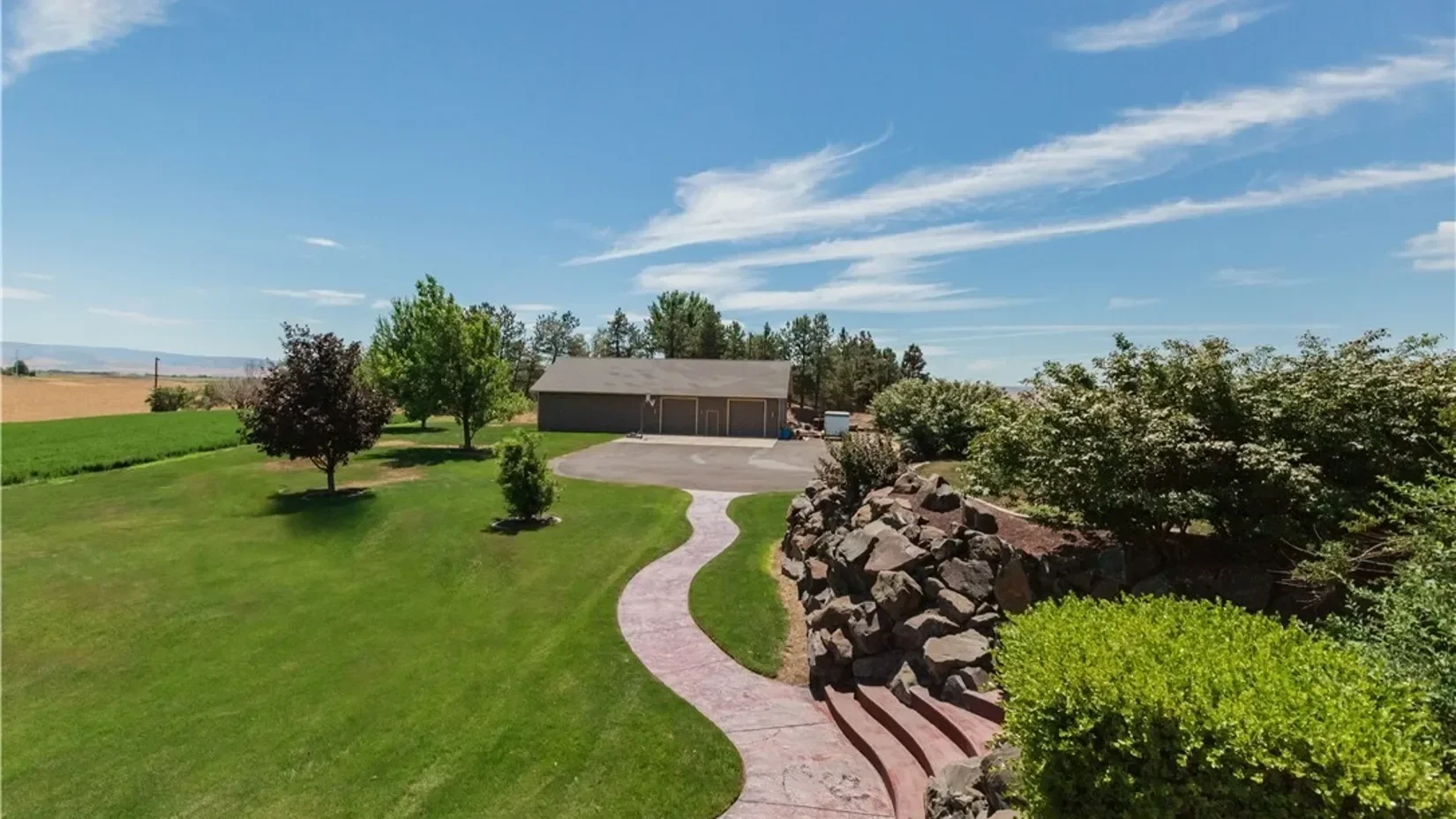 Backyard with a curved concrete pathway, green grass, various trees, rocks, and a storage shed under a blue sky with white clouds.