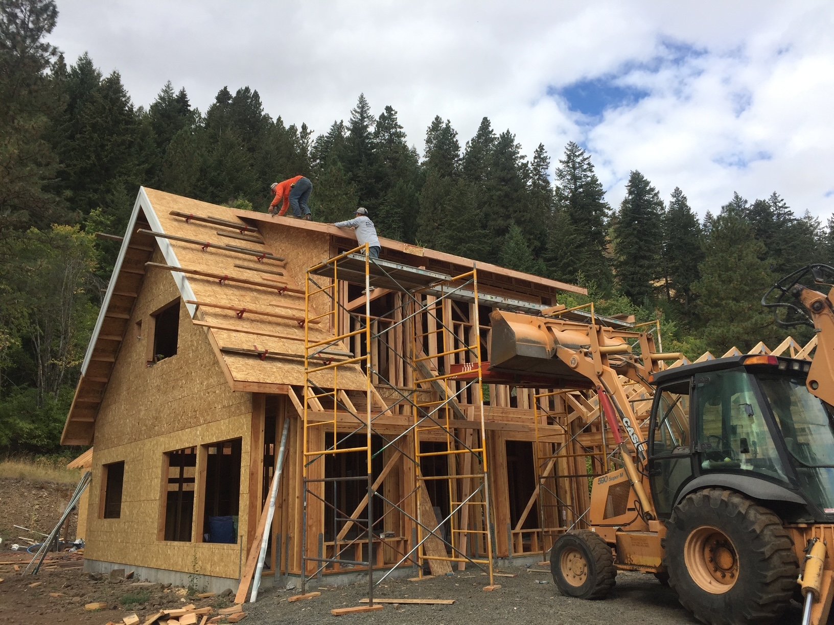 Construction workers on the roof of a wooden house under construction, with scaffolding and a backhoe loader in front, surrounded by trees and a cloudy sky.