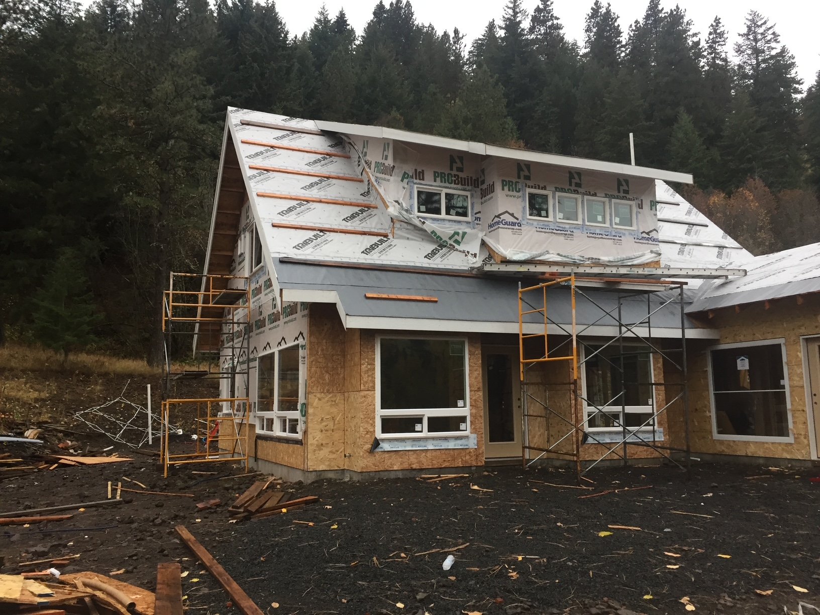 A house under construction with scaffolding, partially covered in building wrap, and debris scattered on the ground, surrounded by trees.