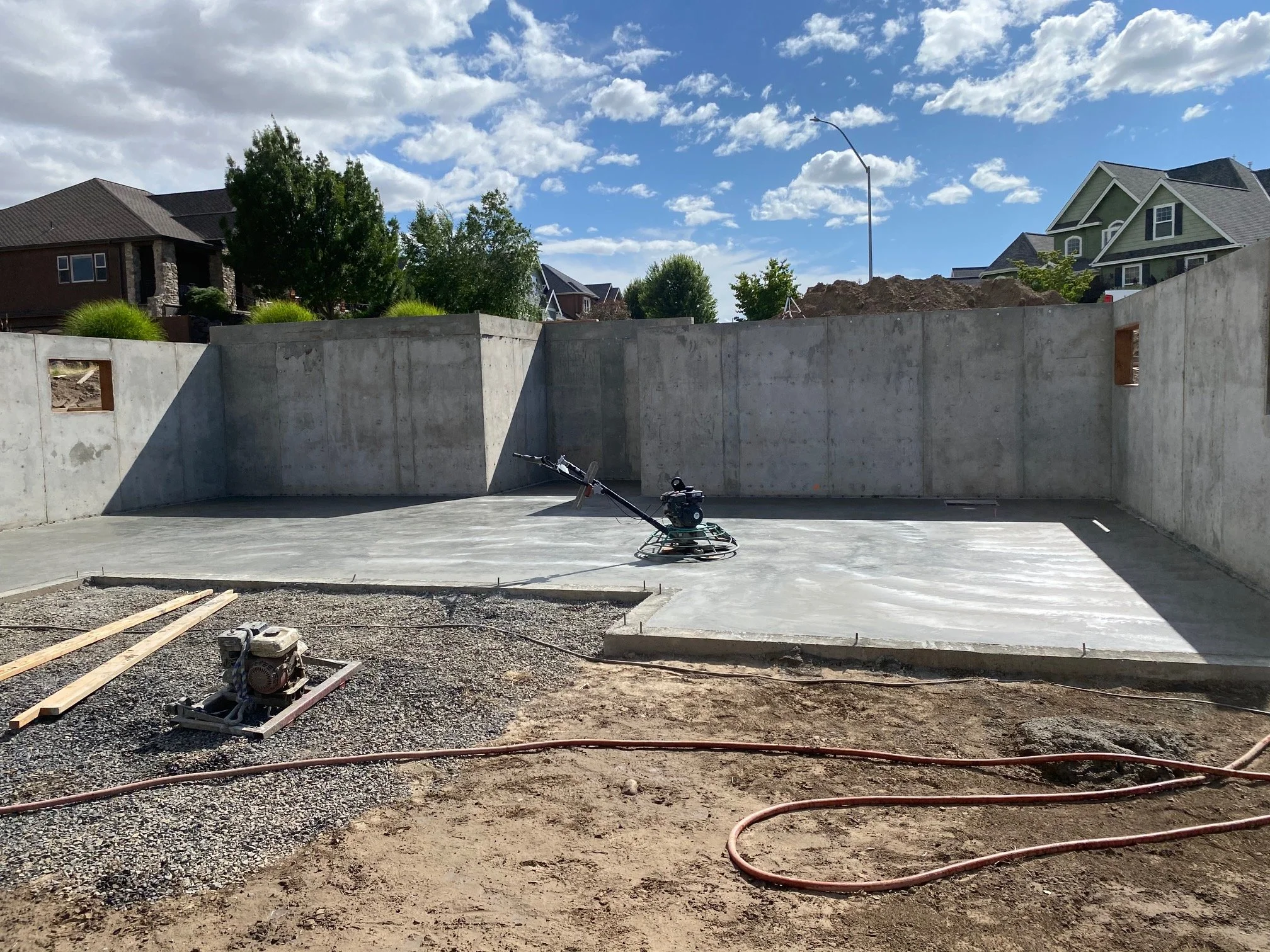 Construction site with concrete foundation walls, construction tools, and a scooter, surrounded by houses and trees under a cloudy sky.