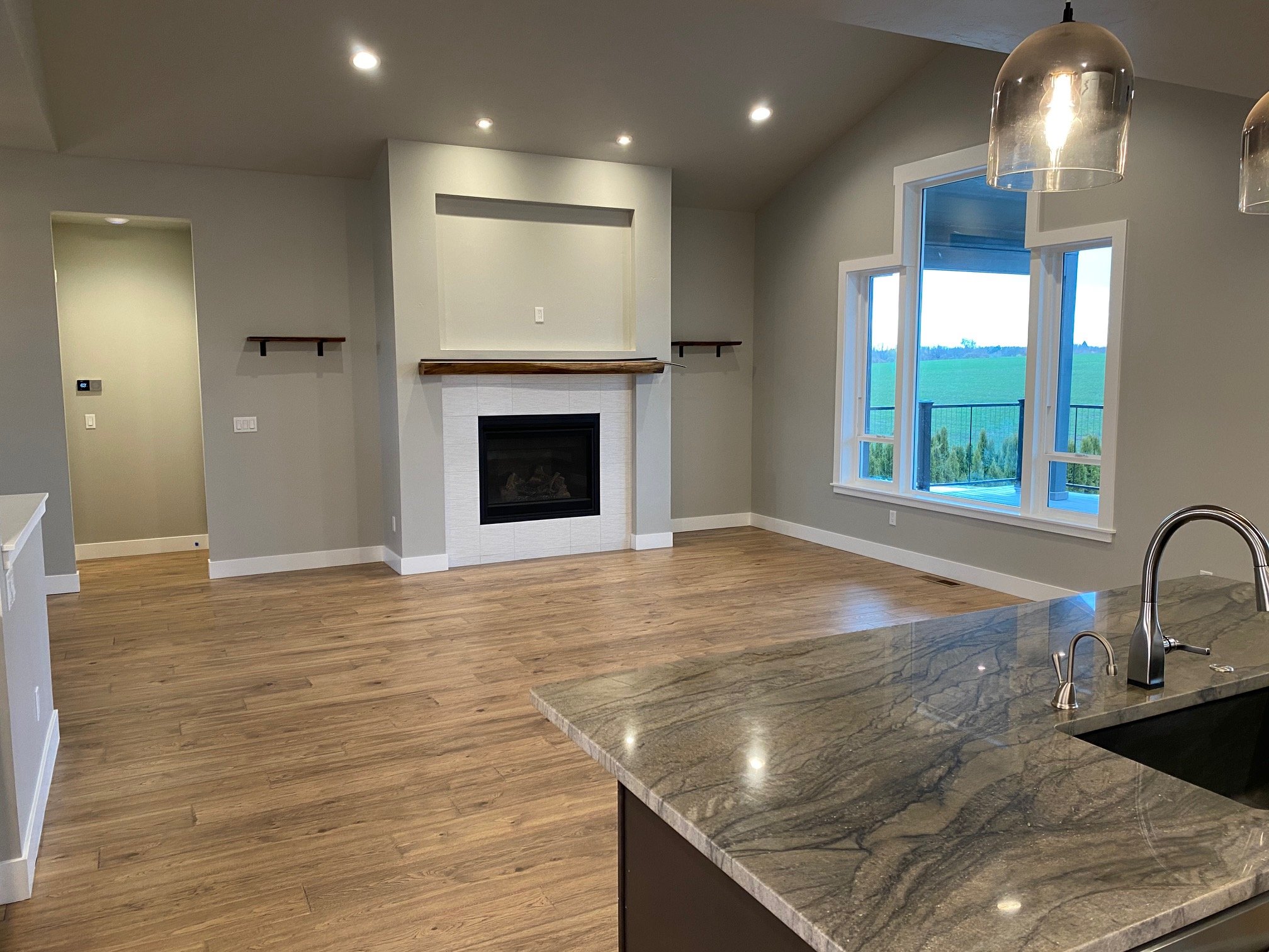 Modern kitchen with a view outside through large windows, hardwood flooring, a fireplace on the wall, and a granite countertop with a stainless steel sink.