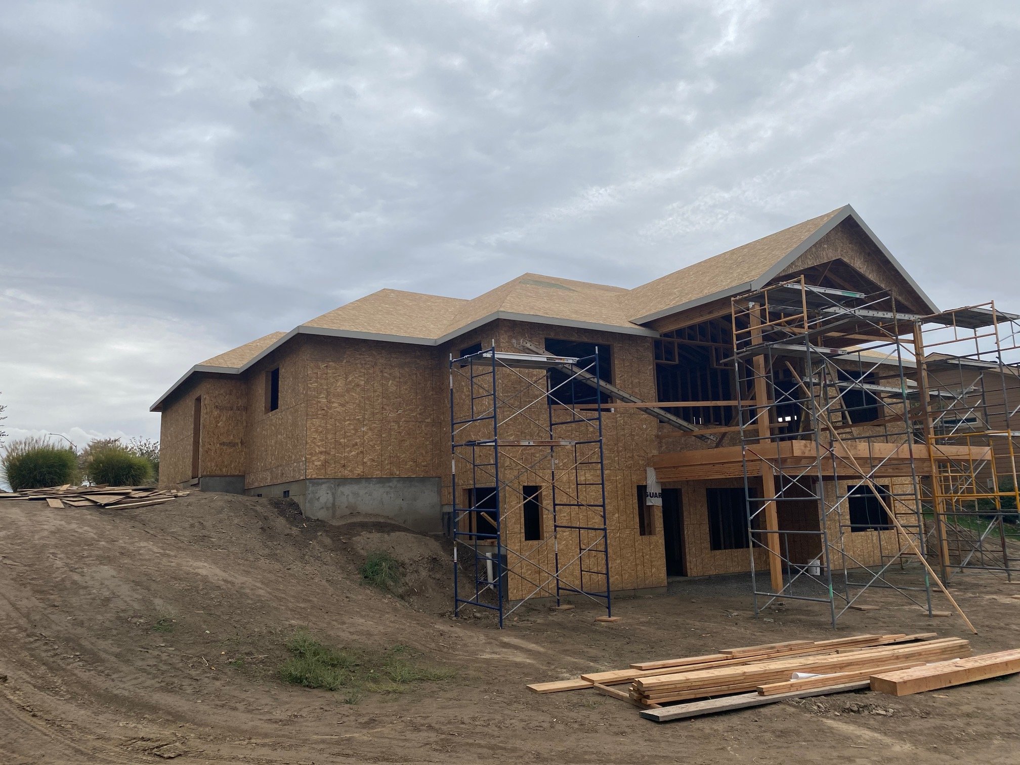 Two-story house under construction with scaffolding, showing an unfinished exterior with exposed wooden sheathing, a pitched roof, and open window spaces; construction materials and wooden planks in the foreground; a dirt pathway leading up to the ho