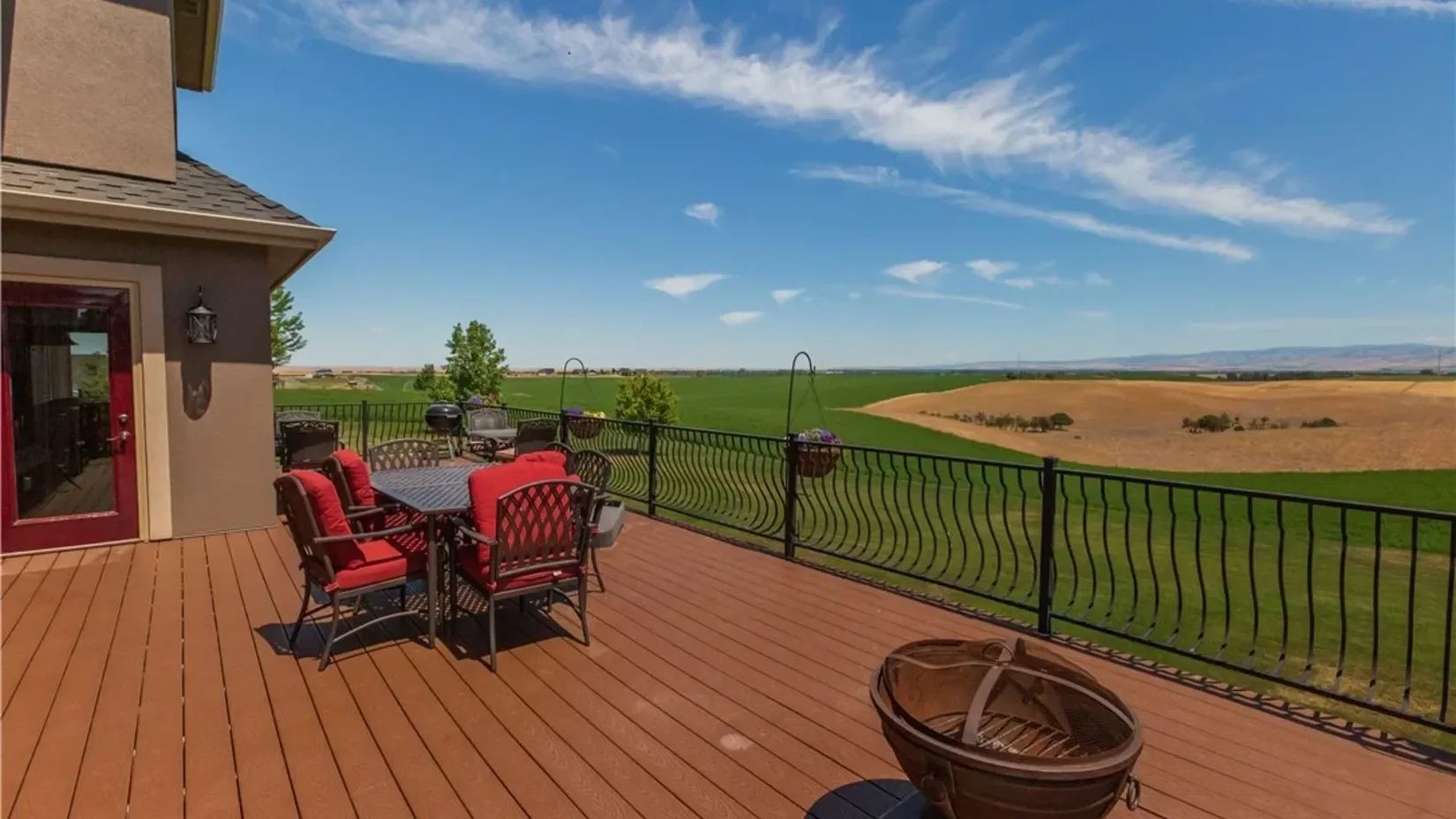 Outdoor wooden deck with a dining table and chairs with red cushions, overlooking green fields and rolling hills under a blue sky with scattered clouds.