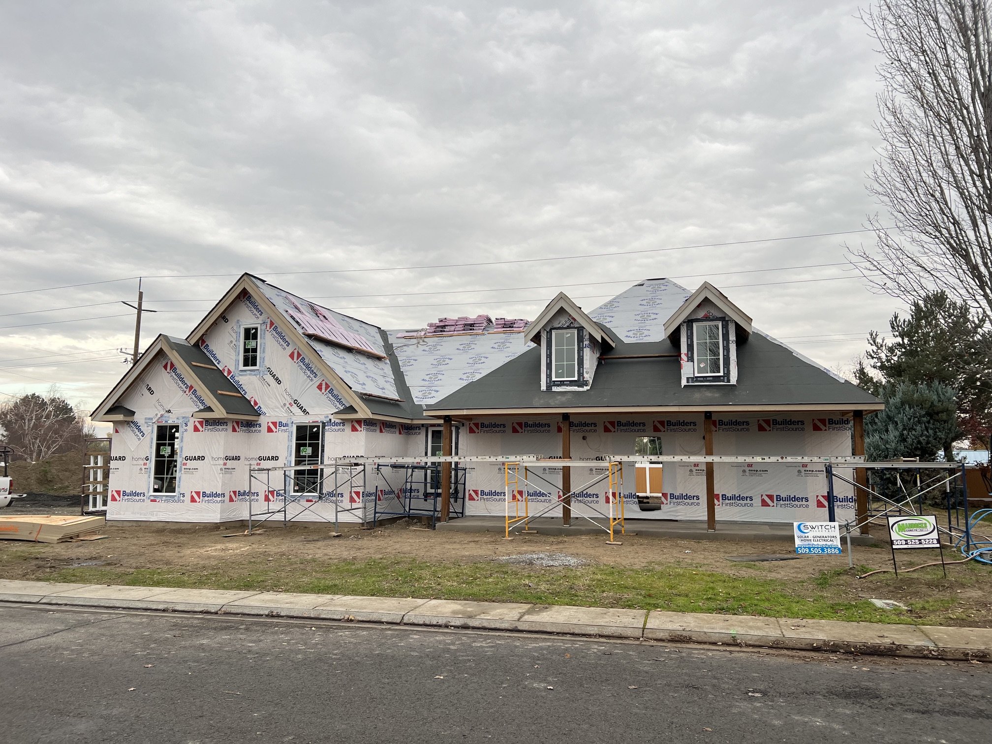 A house under construction with partially installed roofing, window frames, and scaffolding, surrounded by yard and trees, under an overcast sky.