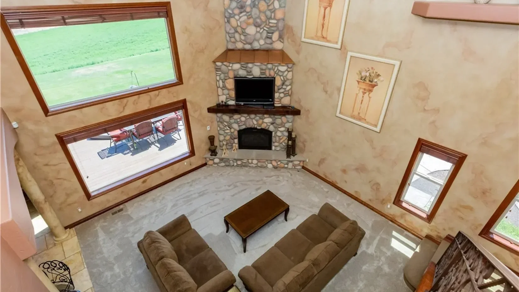 Living room with large windows, a stone fireplace with a TV on top, two brown sofas, a wooden coffee table, and framed artwork on the walls.