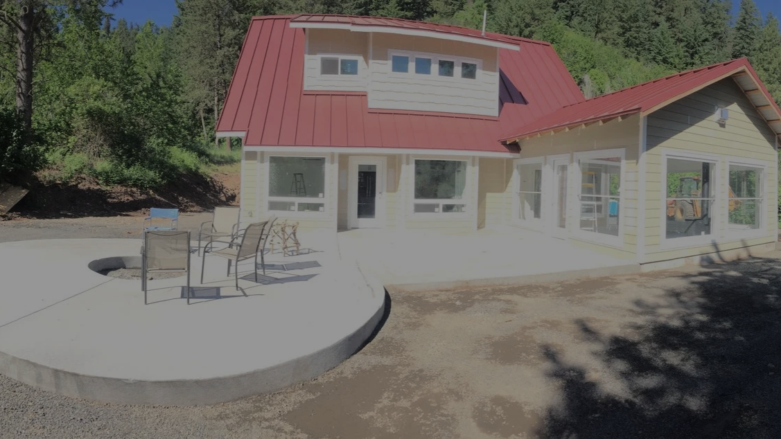New two-story house with a red metal roof, beige siding, and large windows, outdoor patio with chairs and a fire pit, surrounded by trees.